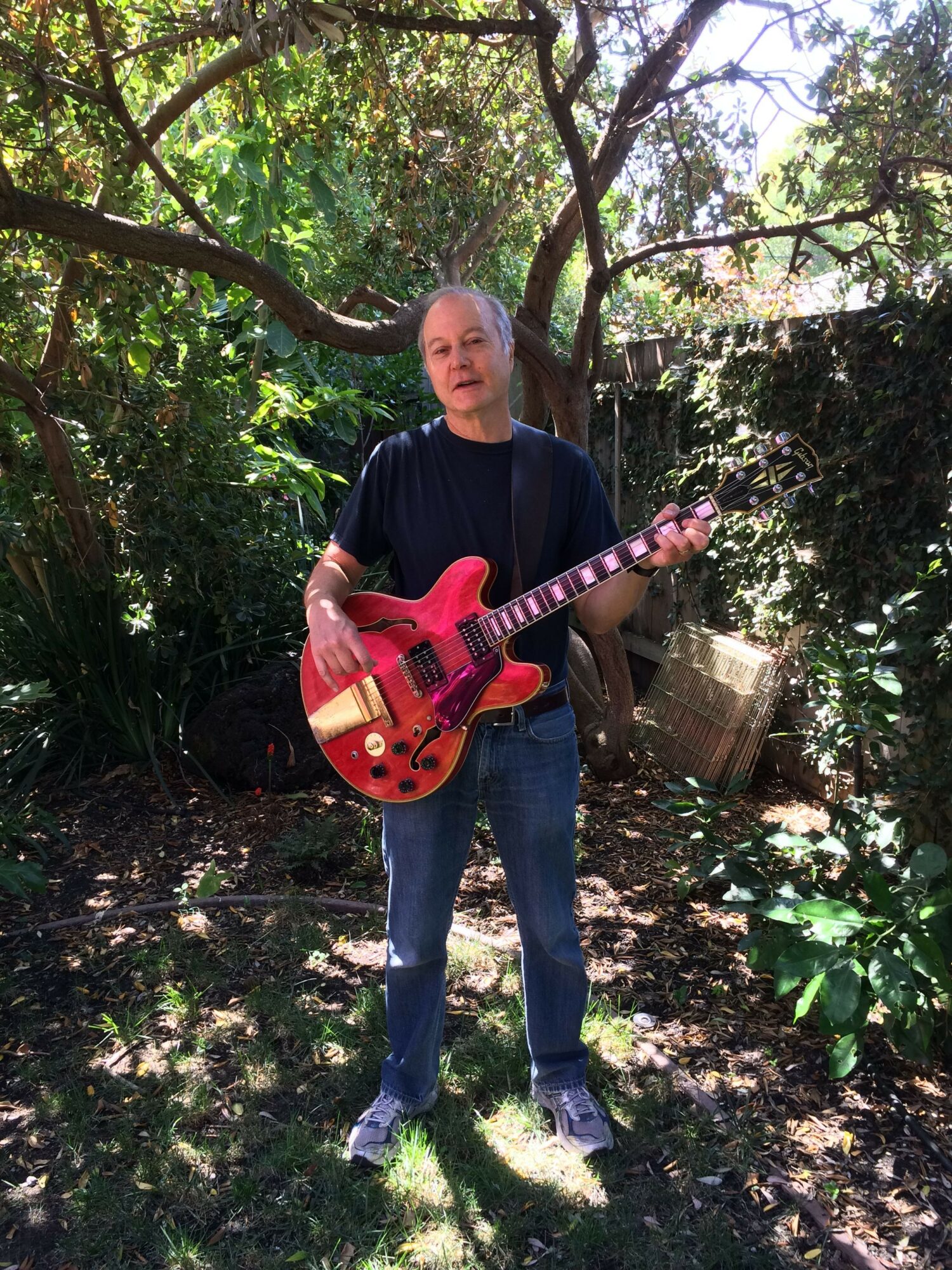 Person standing outdoors holding a red electric guitar, surrounded by trees and greenery.