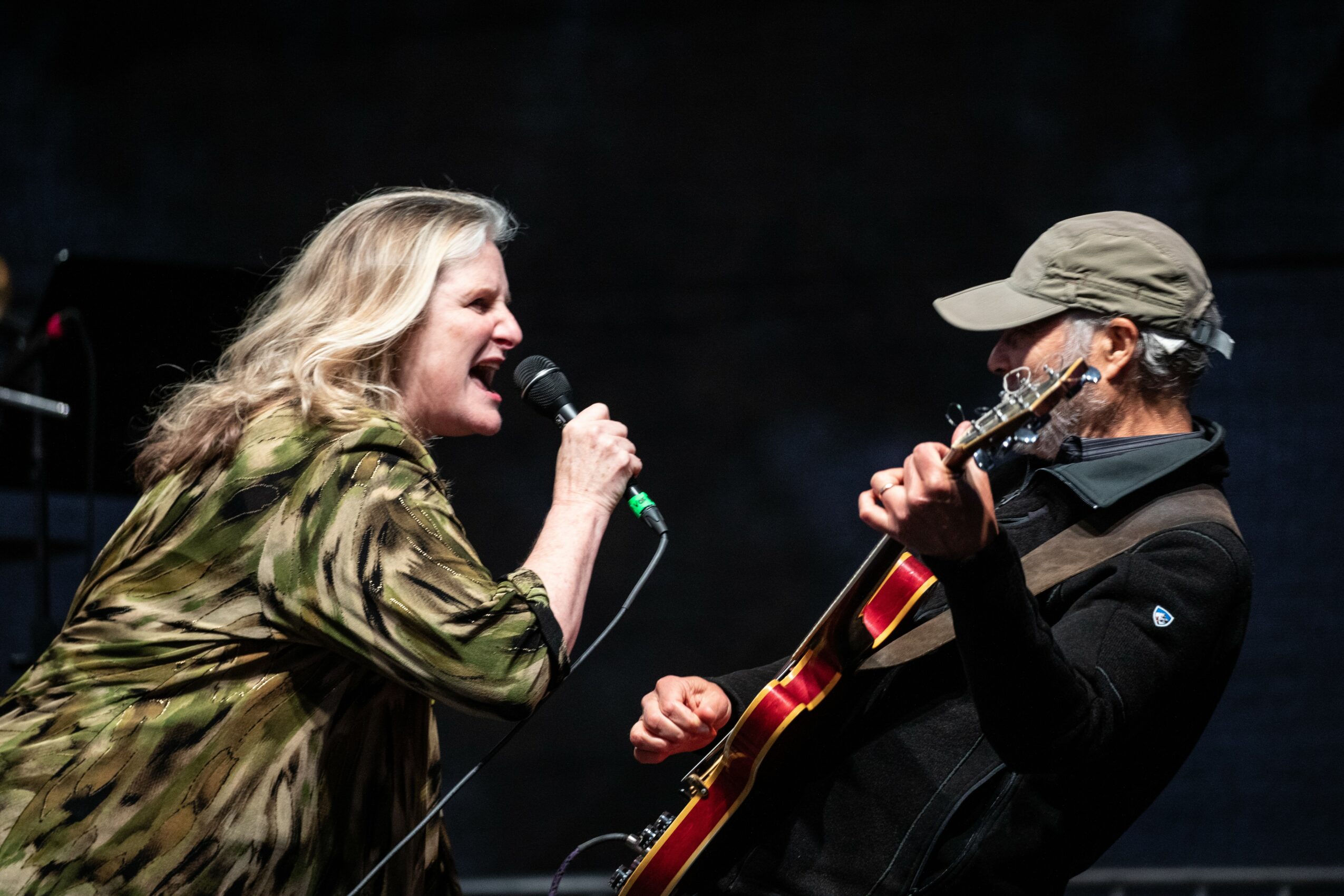 Two people perform on stage; one sings into a microphone, the other plays a guitar, both engaged in music.