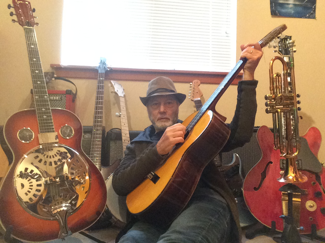 Man wearing a hat playing an acoustic guitar in a room with various guitars on the wall behind him.