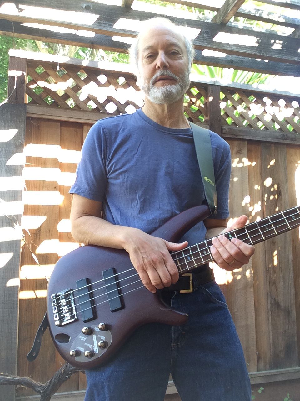 Older man with gray hair and beard holding a dark electric guitar outdoors, wooden fence background.