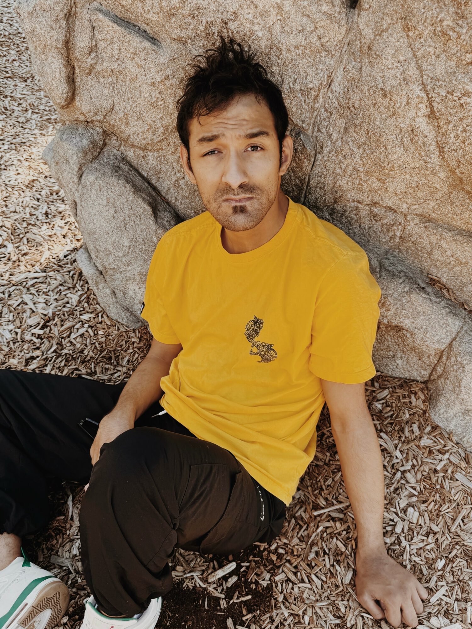 Man sitting on ground against rocks, wearing yellow T-shirt and black pants, looking at camera.