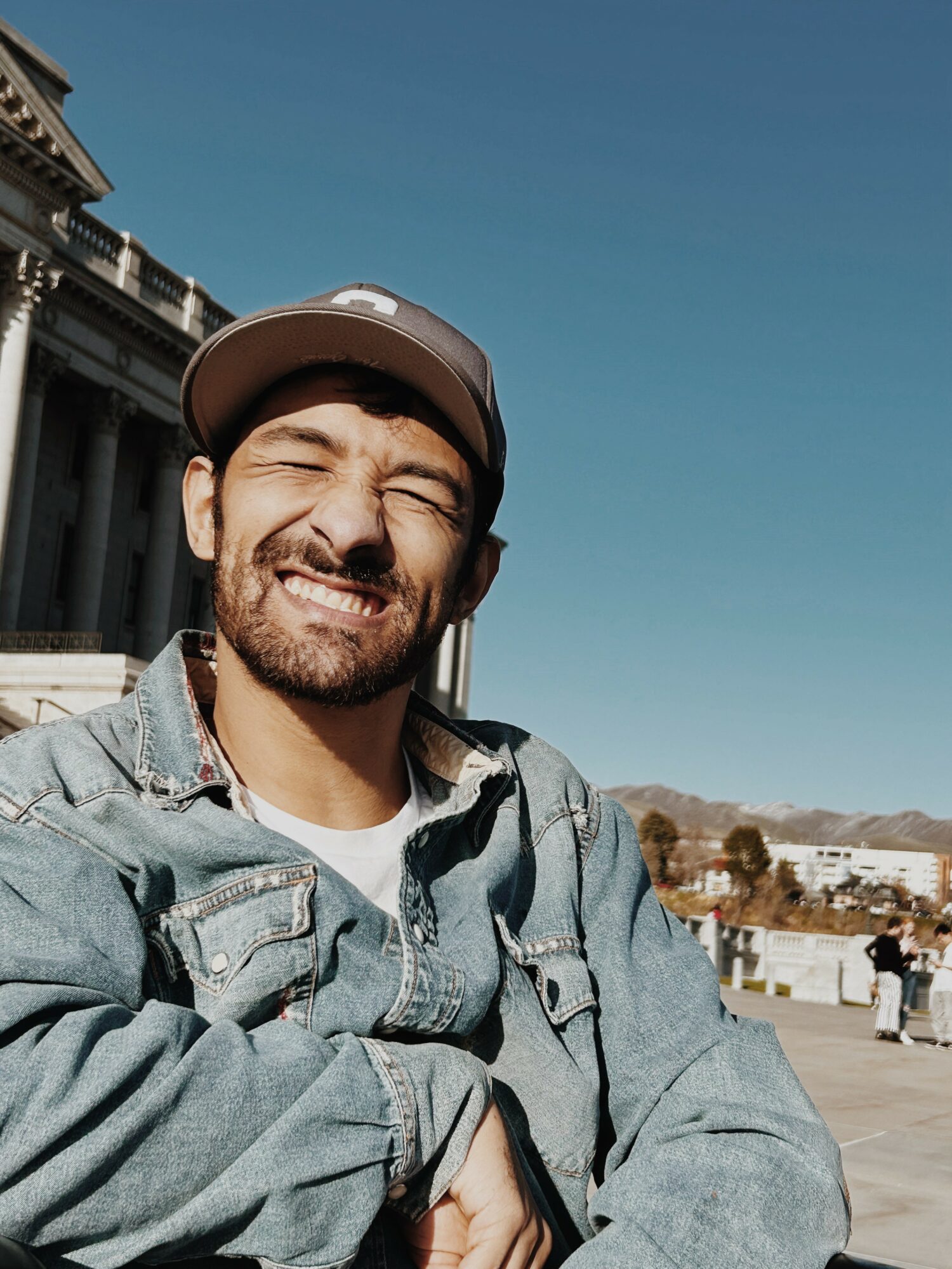 Man with a beard wearing a cap and denim jacket smiling outdoors against a clear sky.