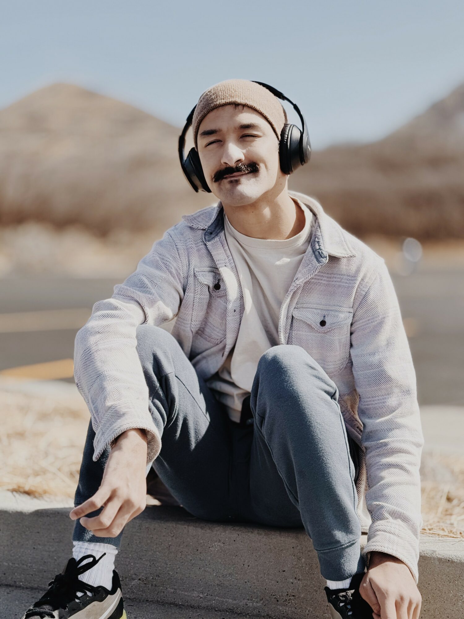 Young man sitting outdoors, wearing headphones, a beanie, and casual clothing, smiling with a desert landscape background.