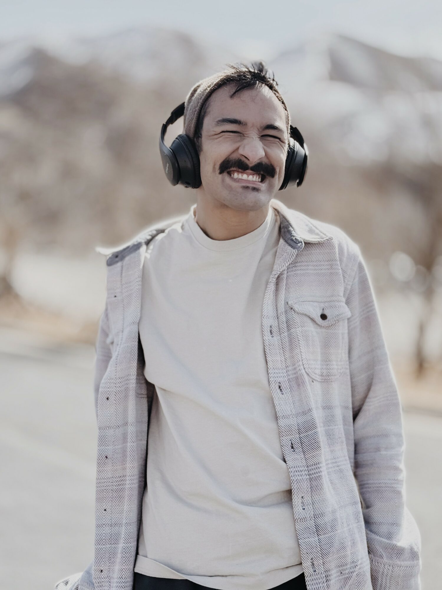 Man with headphones smiling outdoors, wearing a light shirt and jacket, with blurred trees in background.