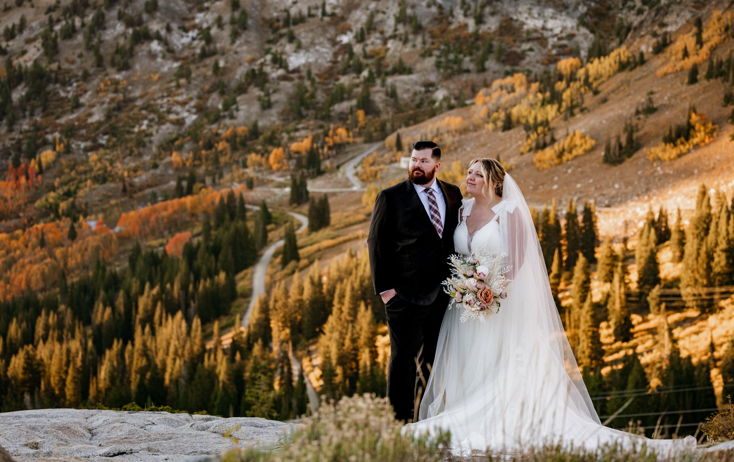 A bride and groom stand outdoors on a rocky area with a forested mountain landscape in the background.