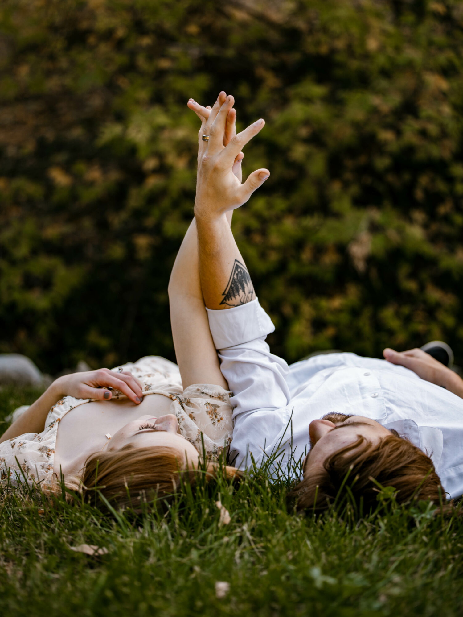 Person lying on grass with arm raised, tattoo visible, outdoors with blurred green foliage background.