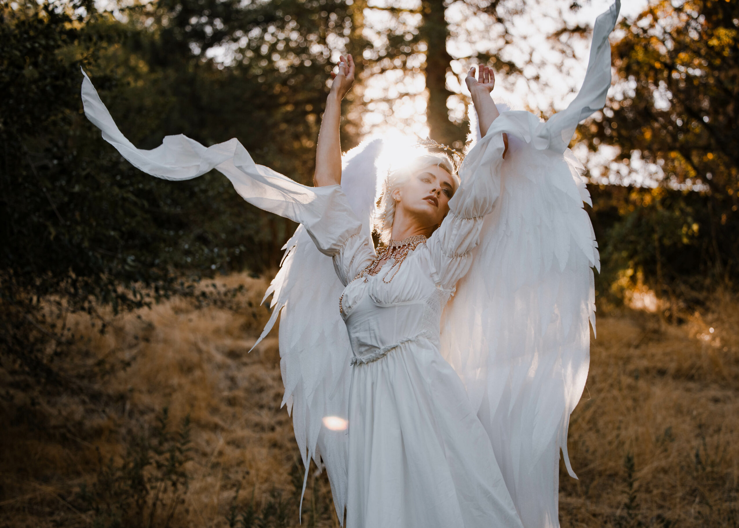 Woman dressed as an angel with large white wings outdoors, sunlight shining through trees behind her.