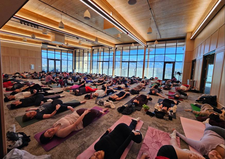 Large group of people practicing yoga or meditation on mats in a spacious room with floor-to-ceiling windows and wooden ceiling.