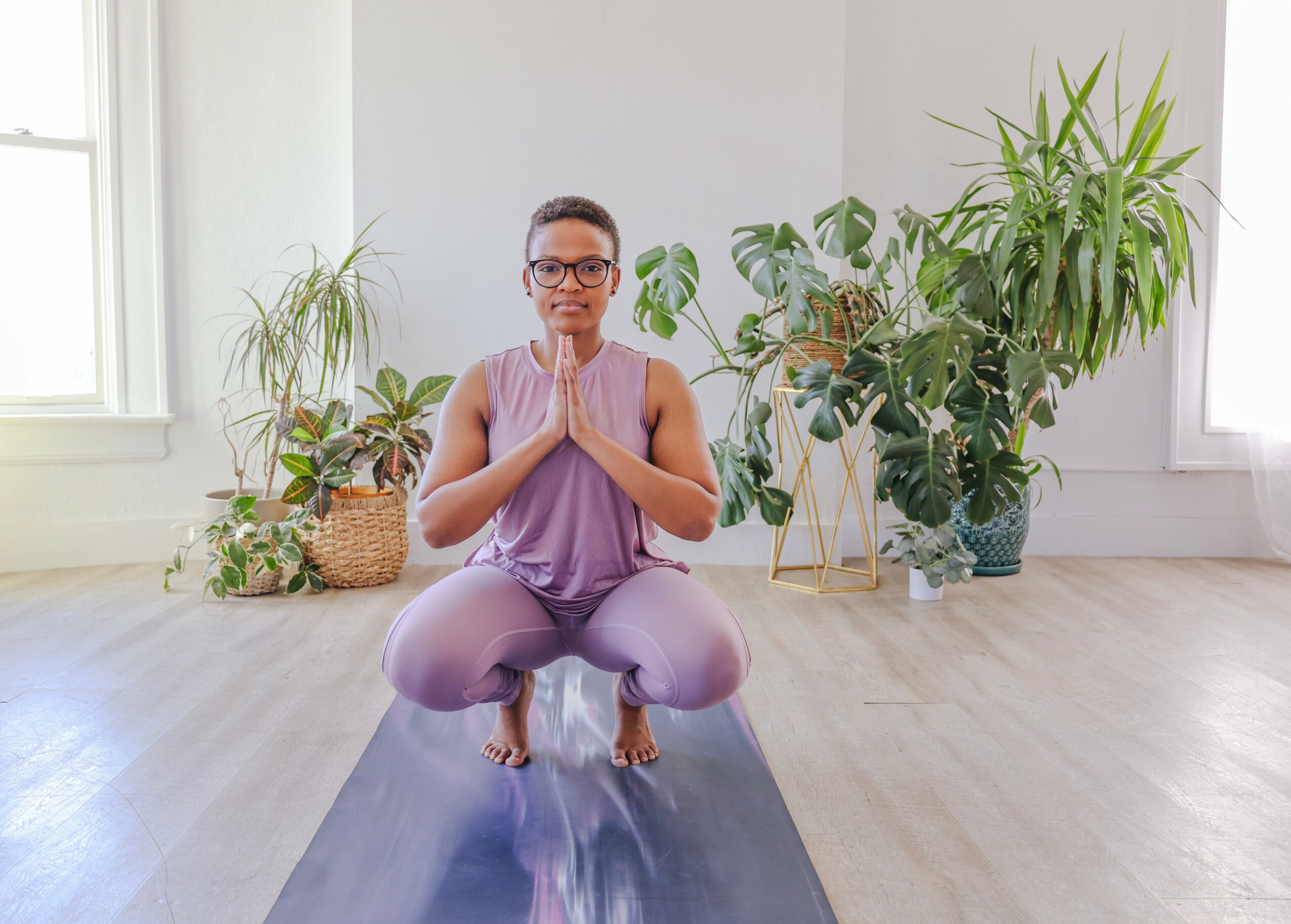 Person practicing yoga on a mat with hands in prayer position, surrounded by potted plants in a bright room.