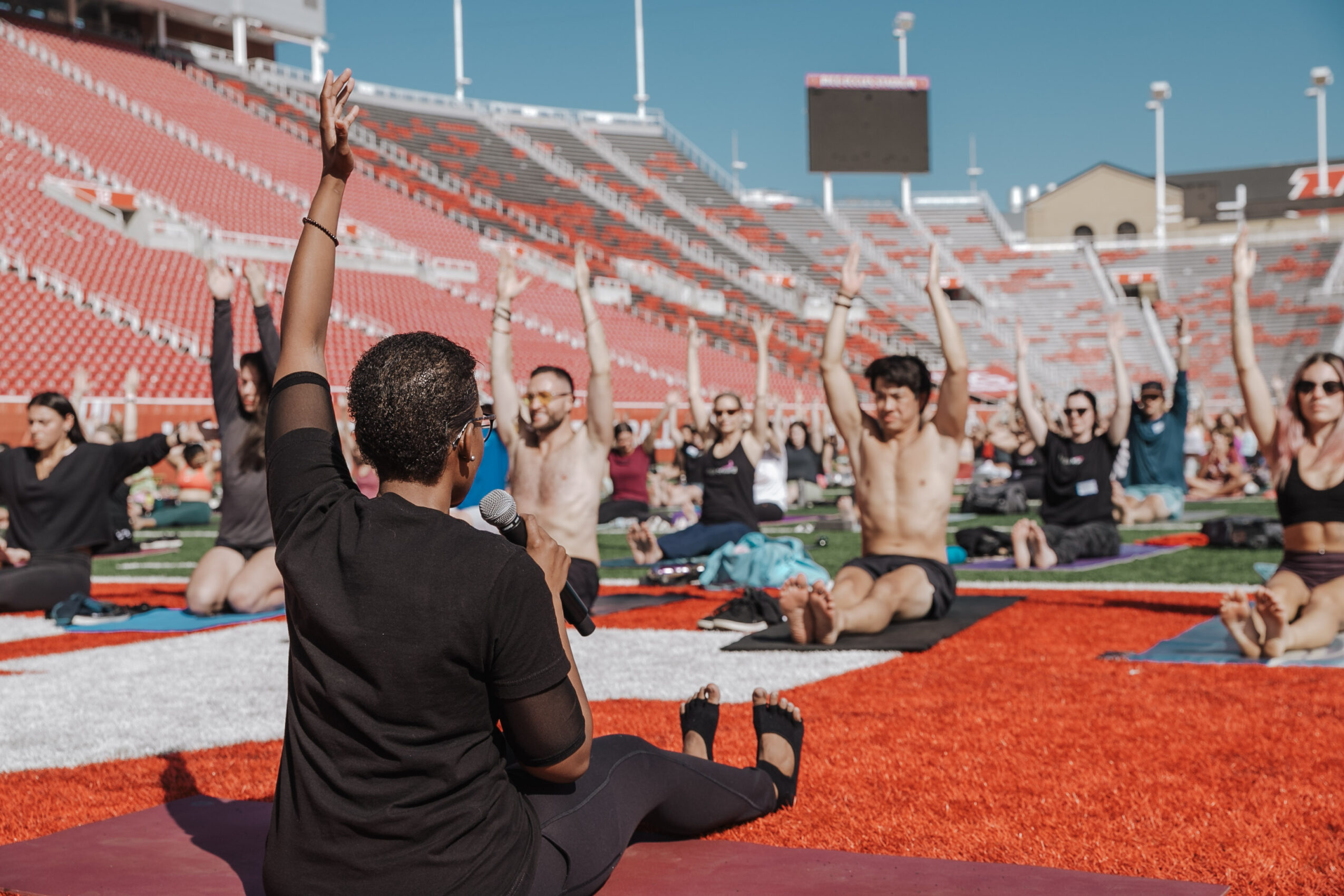 Group of people practicing yoga outdoors on mats in a stadium, raising arms, with empty red seats in background.
