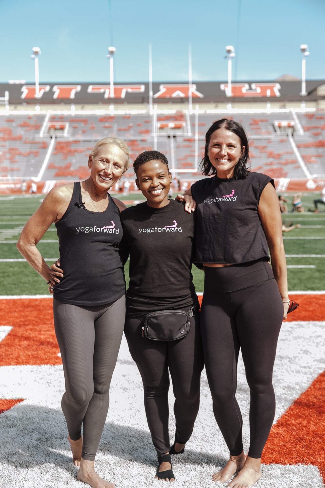 Three women standing together on a sports field with stadium seats in the background.