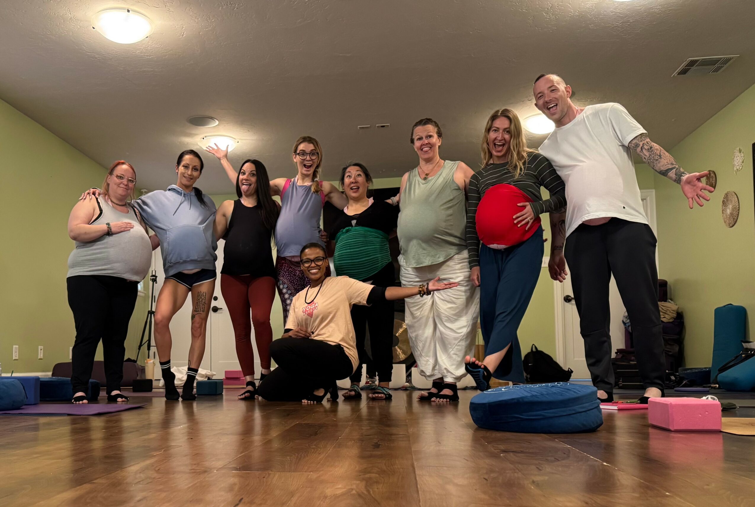 Group of ten people standing in a room, smiling, some holding exercise balls, with yoga mats on the floor.