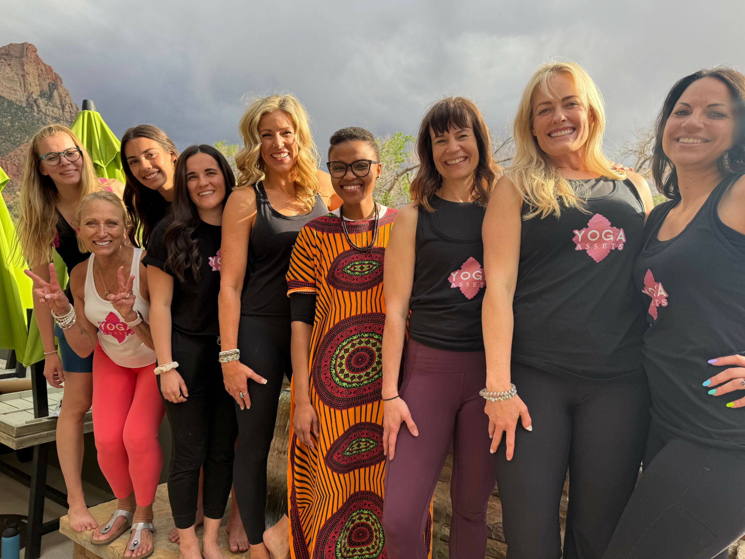 Group of women standing outdoors, smiling, with mountains and cloudy sky in background.