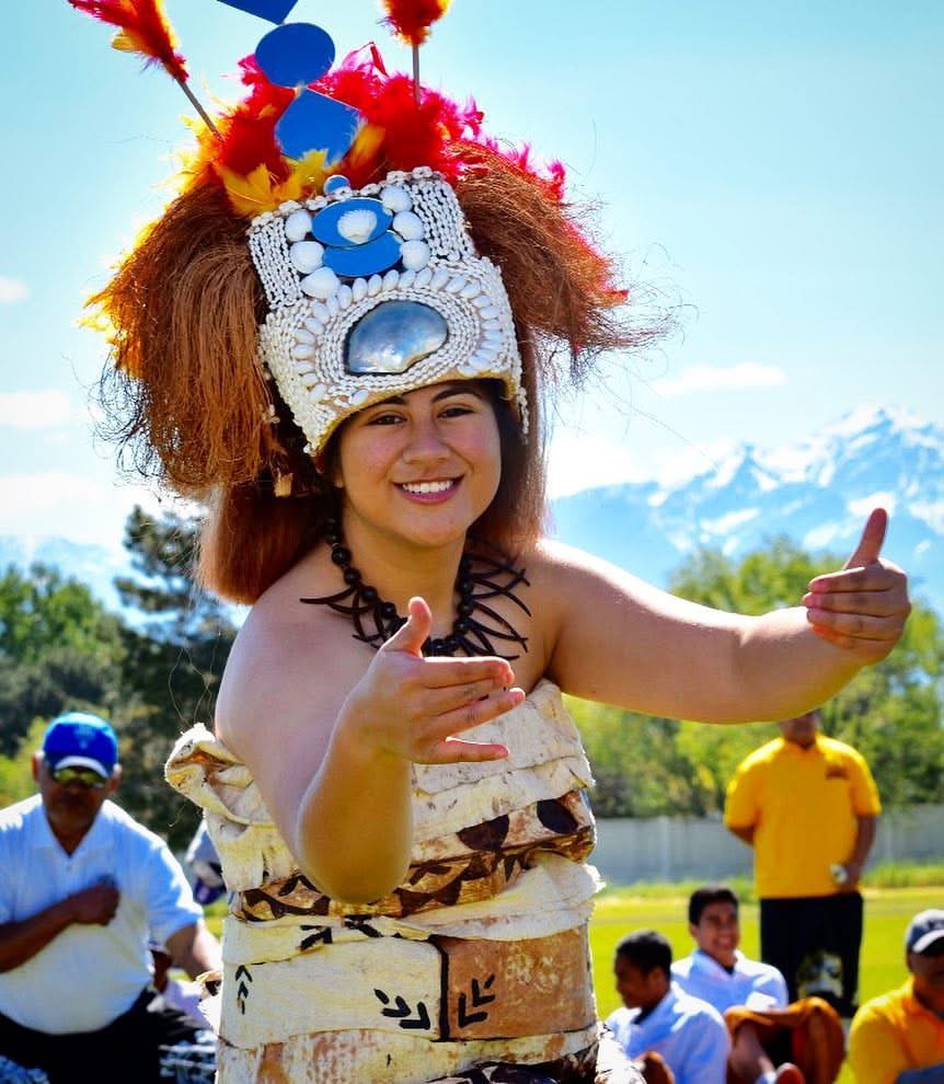 Child wearing a colorful headdress and traditional clothing, smiling outdoors with mountains and trees in background.