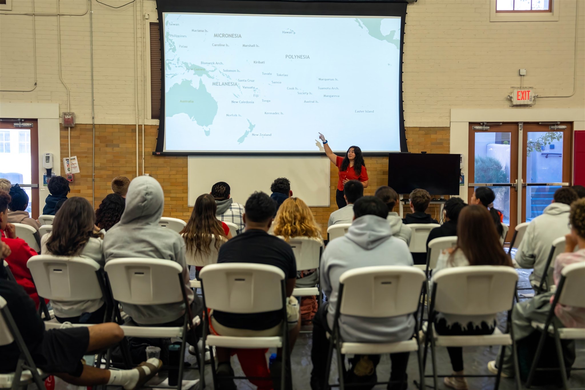 A woman presents in front of a large screen with a map, audience seated in chairs facing her in a room.