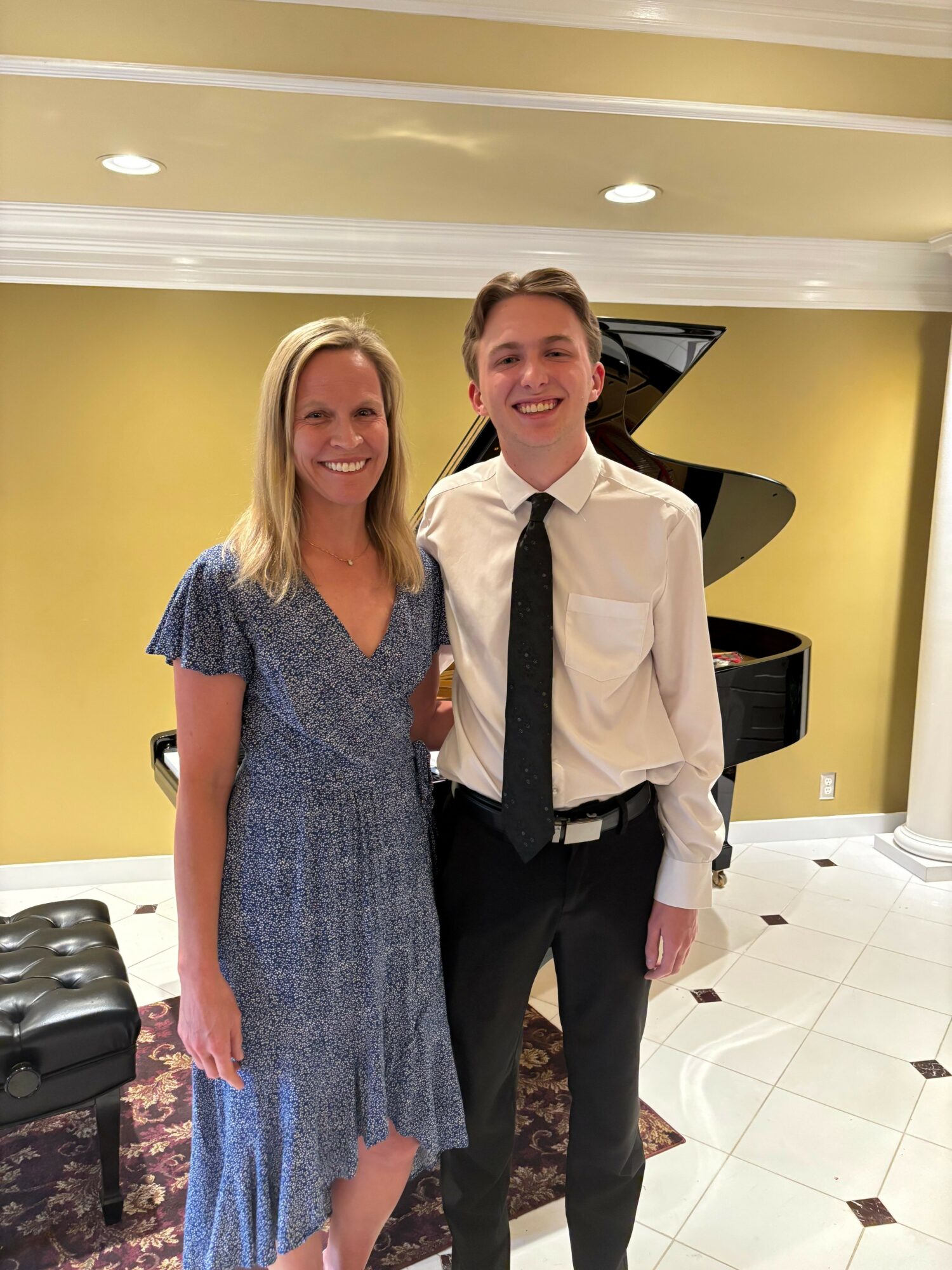 A woman and a young man stand side by side in front of a grand piano, smiling at the camera.