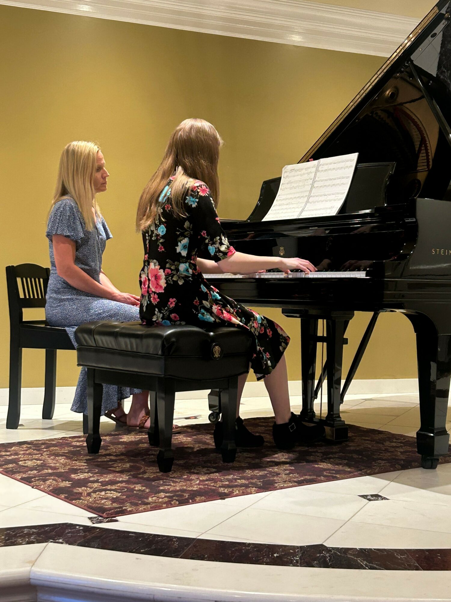 Two girls playing piano in a room with a green wall and a patterned rug.