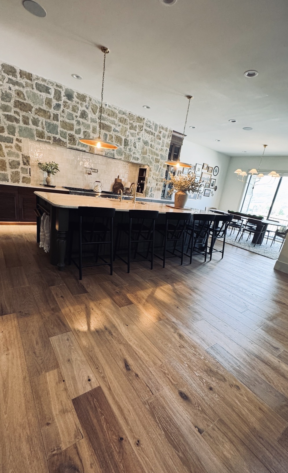 Modern kitchen with a large island, black chairs, stone wall, pendant lights, and wooden flooring.