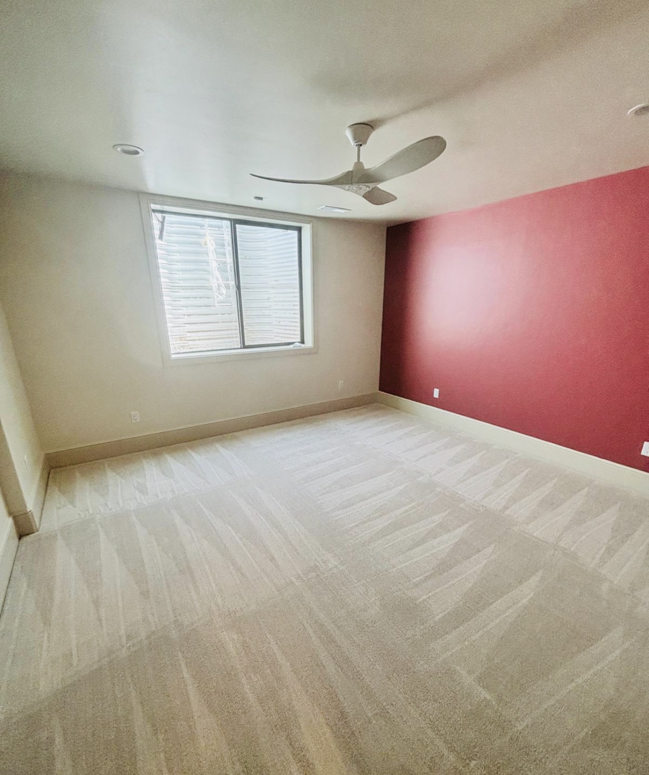 Empty room with beige carpet, white walls, a red accent wall, a window, and a ceiling fan.