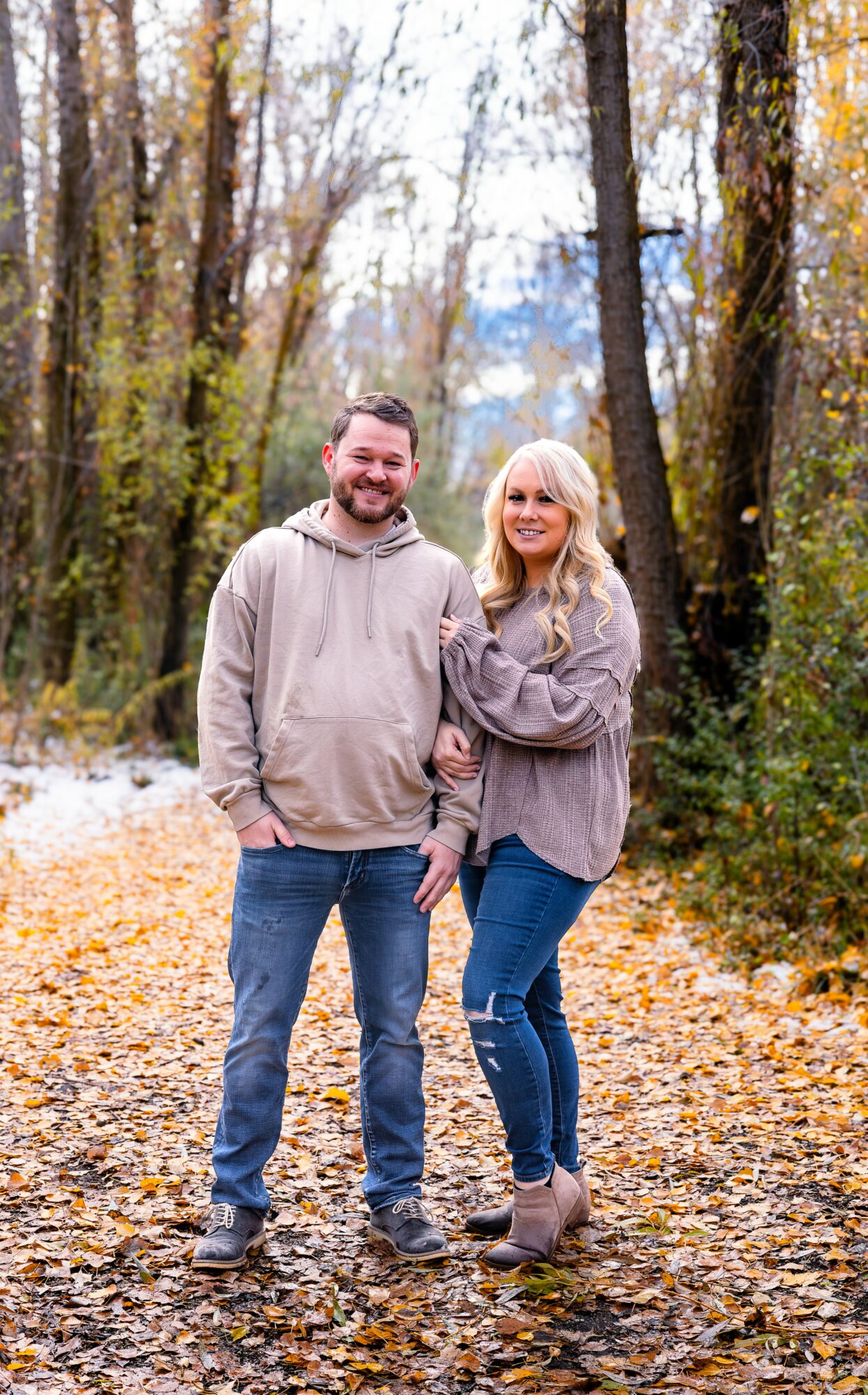 A man and woman stand on a leaf-covered path in a forest during autumn, smiling at the camera.
