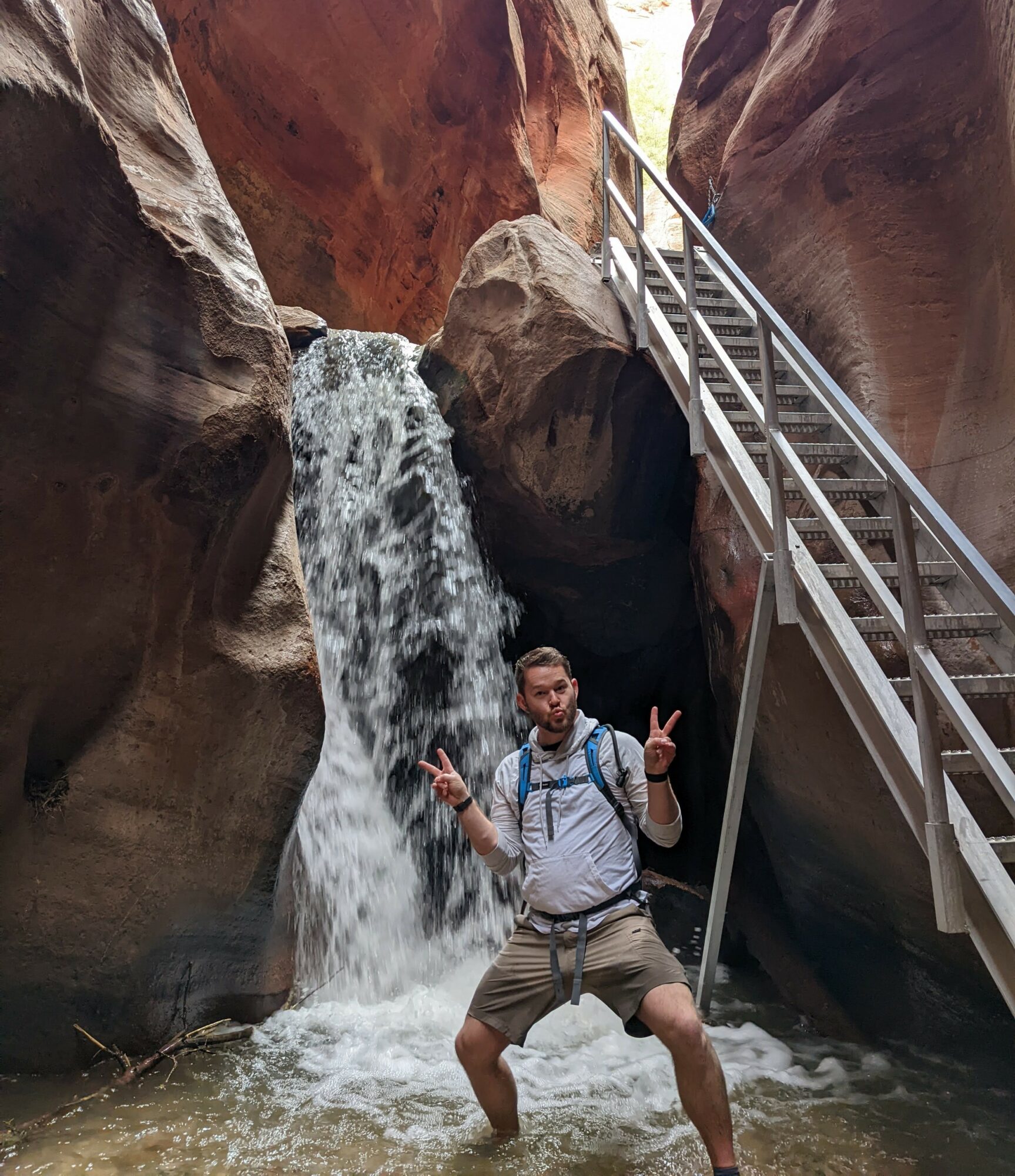 Person standing in a canyon with a waterfall, making peace signs, wearing a backpack and shorts.