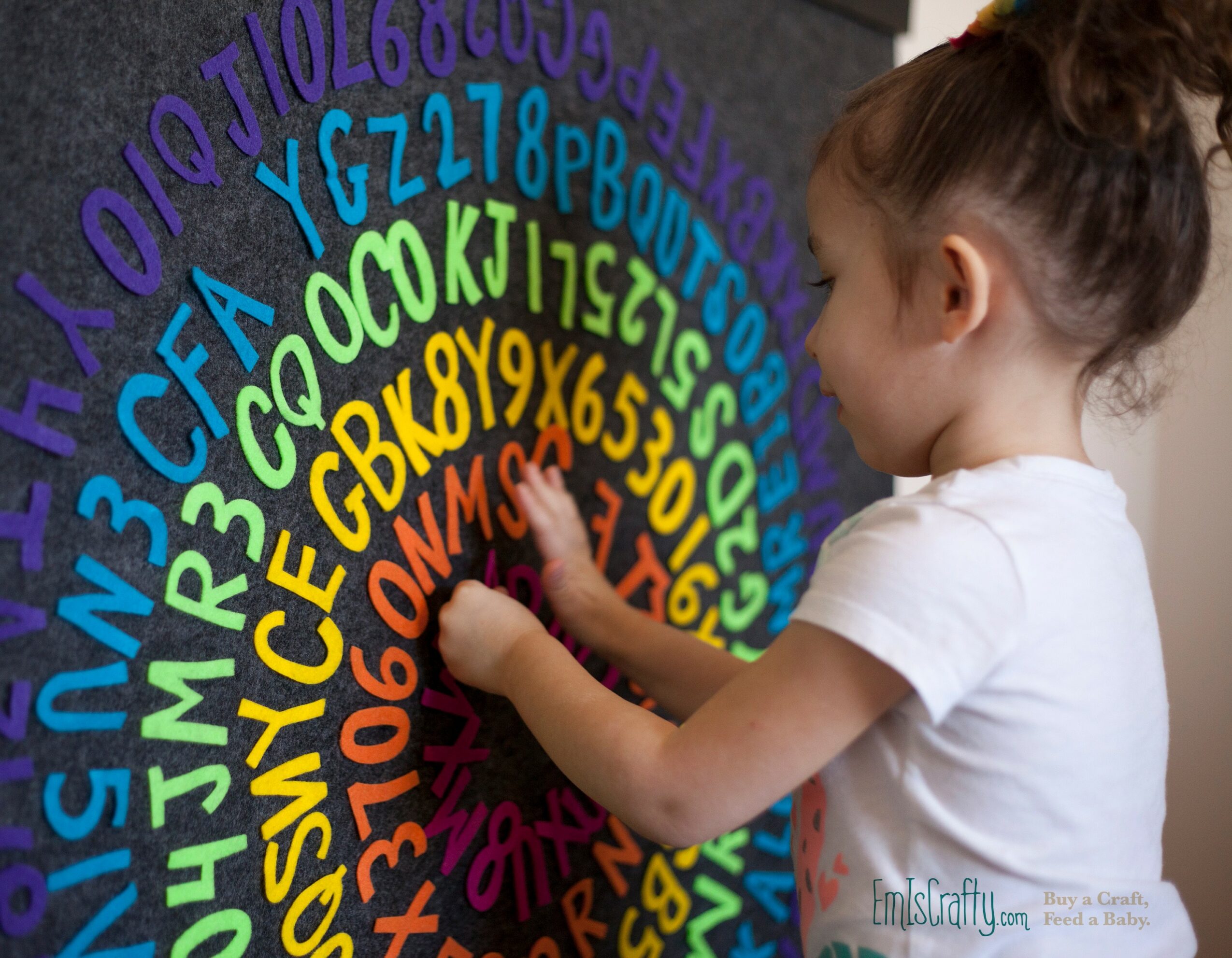Child reaching out to colorful circular alphabet and number display on wall, with hair tied back, wearing a white shirt.