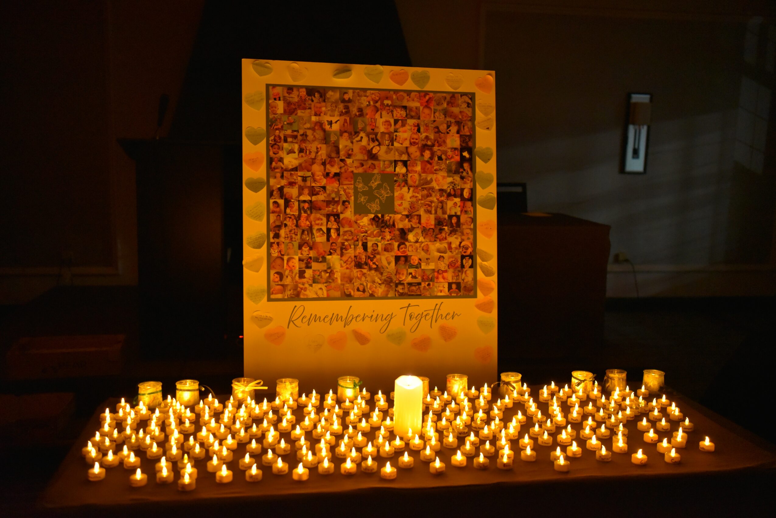 A large illuminated poster with a colorful mosaic and text, surrounded by numerous lit candles on a dark surface.