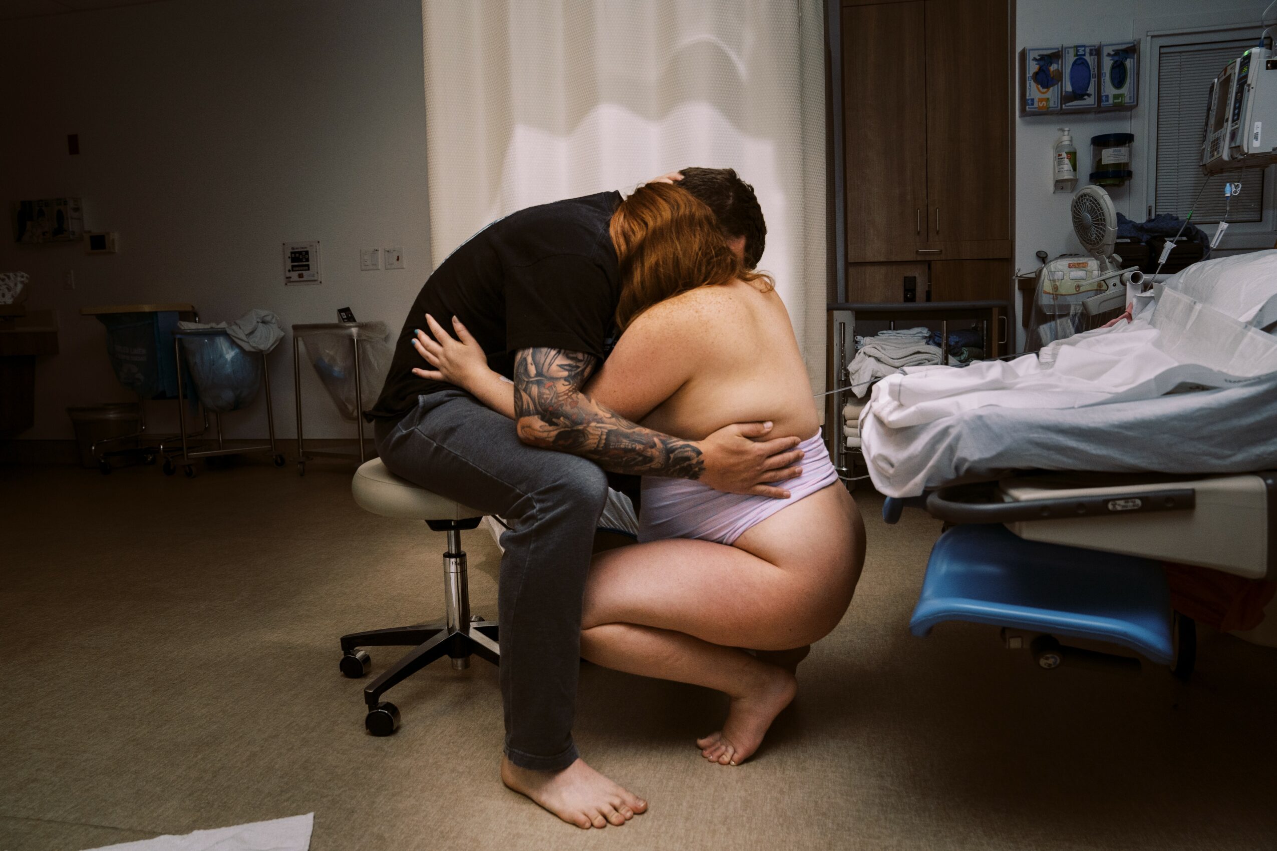 Two people embrace in a hospital room, one sitting on a stool and the other kneeling on the floor, near a hospital bed.