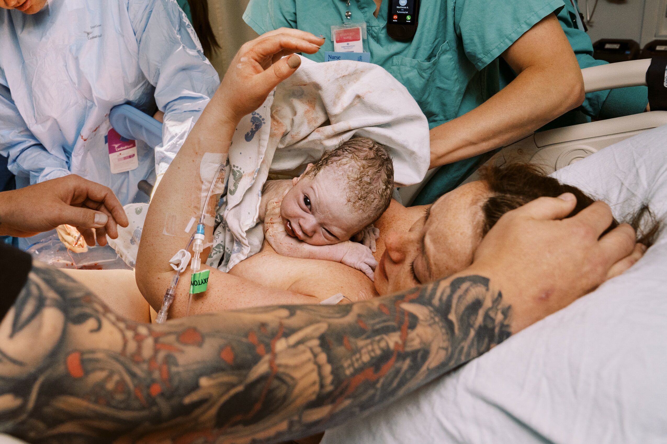 Young boy lying in hospital bed with medical tubes, surrounded by healthcare staff, holding a person's arm with tattoos.