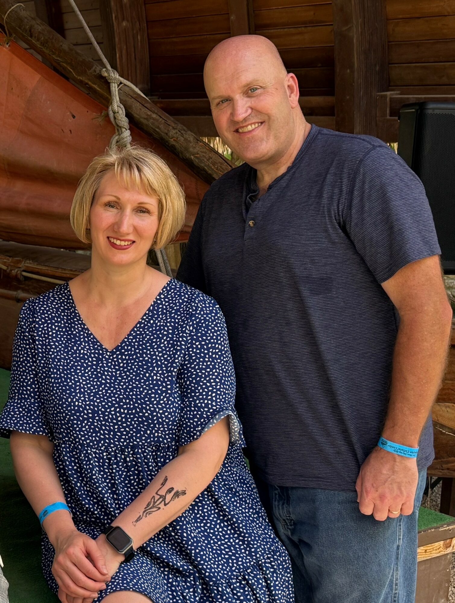 A woman and a man pose together indoors, smiling, with wooden walls and a rope in the background.