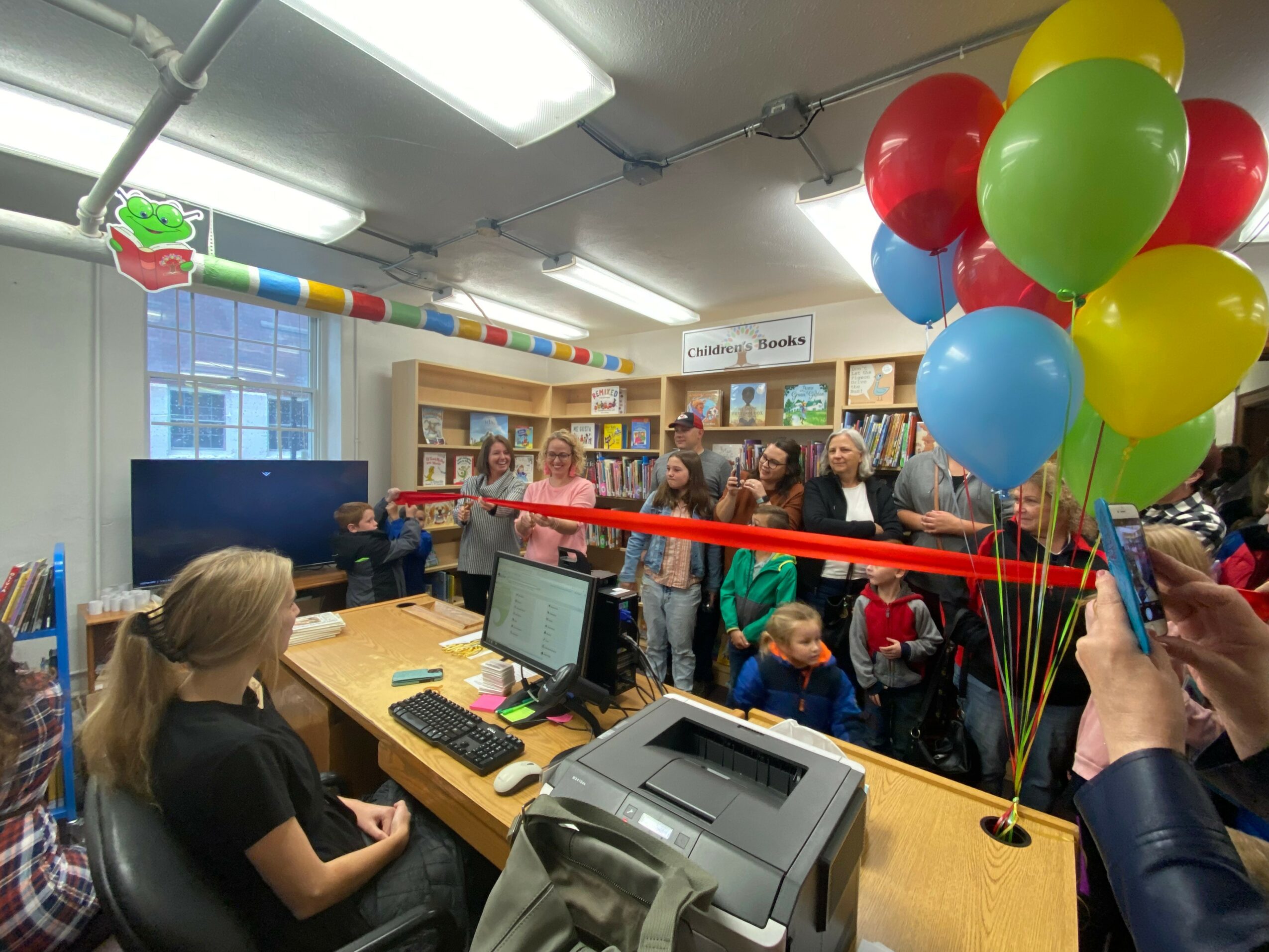 Group of children and adults gathered in a room with bookshelves, celebrating with balloons and a ribbon-cutting ceremony.