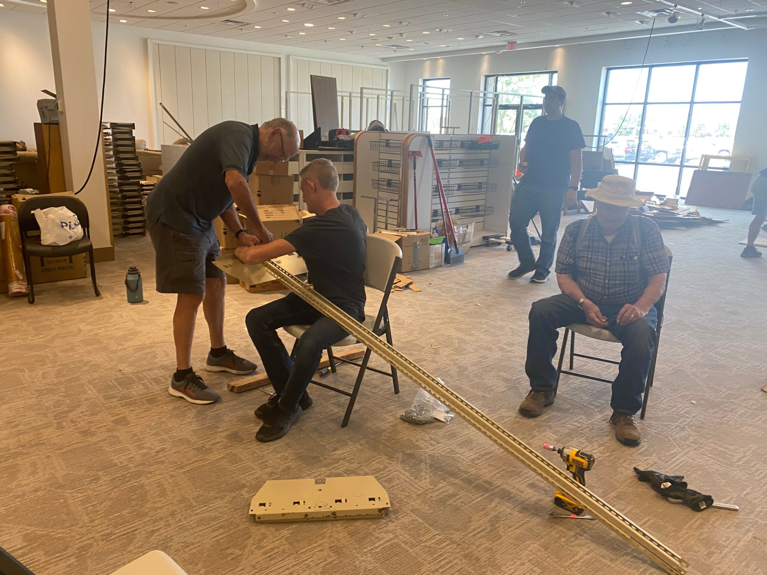 Four people in a room with chairs and shelves, assembling a long wooden structure, with others observing or working nearby.