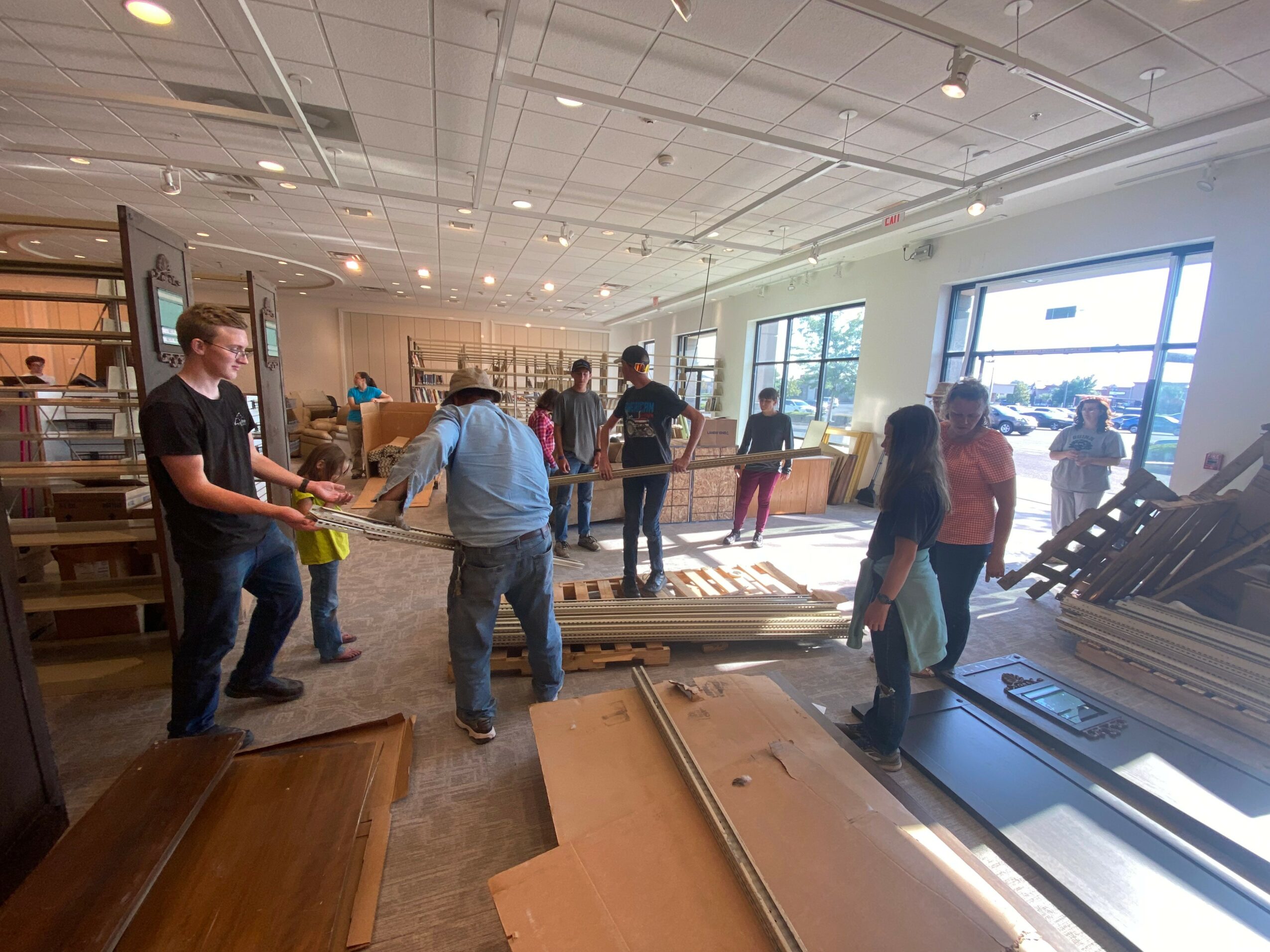 People shopping for furniture in a store, browsing wood and metal items, with shelves and windows in the background.