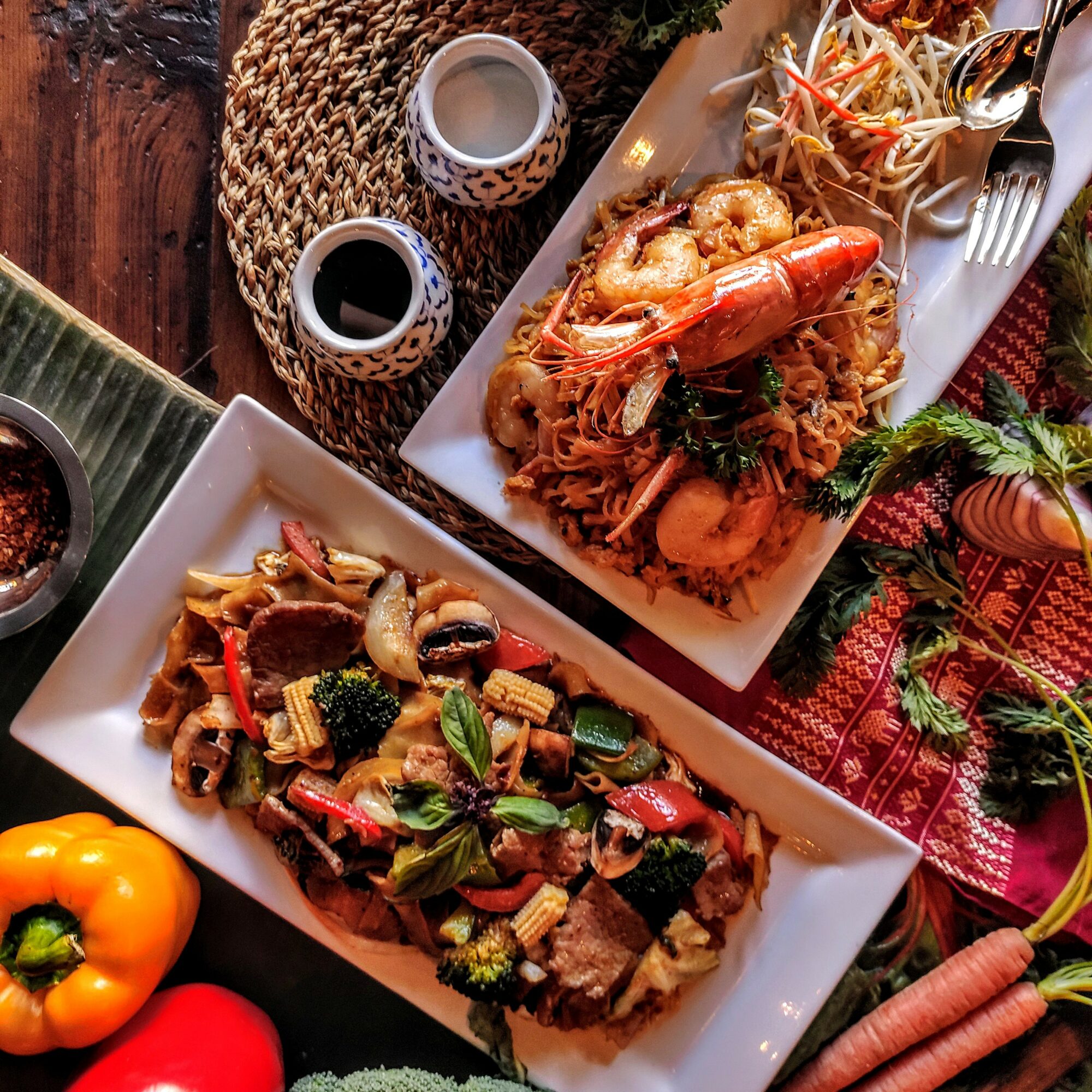 Two rectangular plates with cooked dishes, one with vegetables and meat, the other with seafood, on a table with condiments and vegetables.