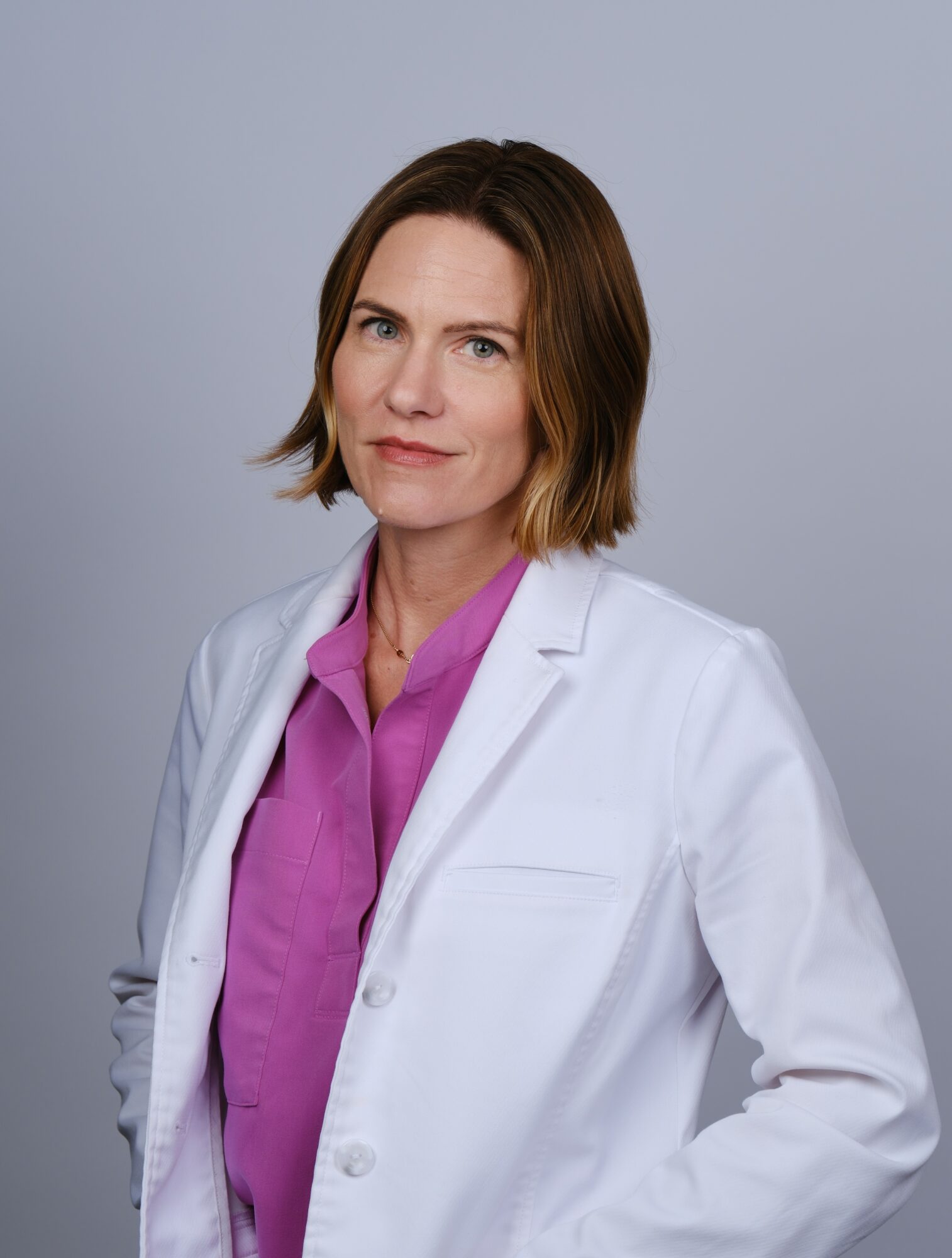 Woman with shoulder-length brown hair wearing a white lab coat and purple shirt, standing against a light gray background.