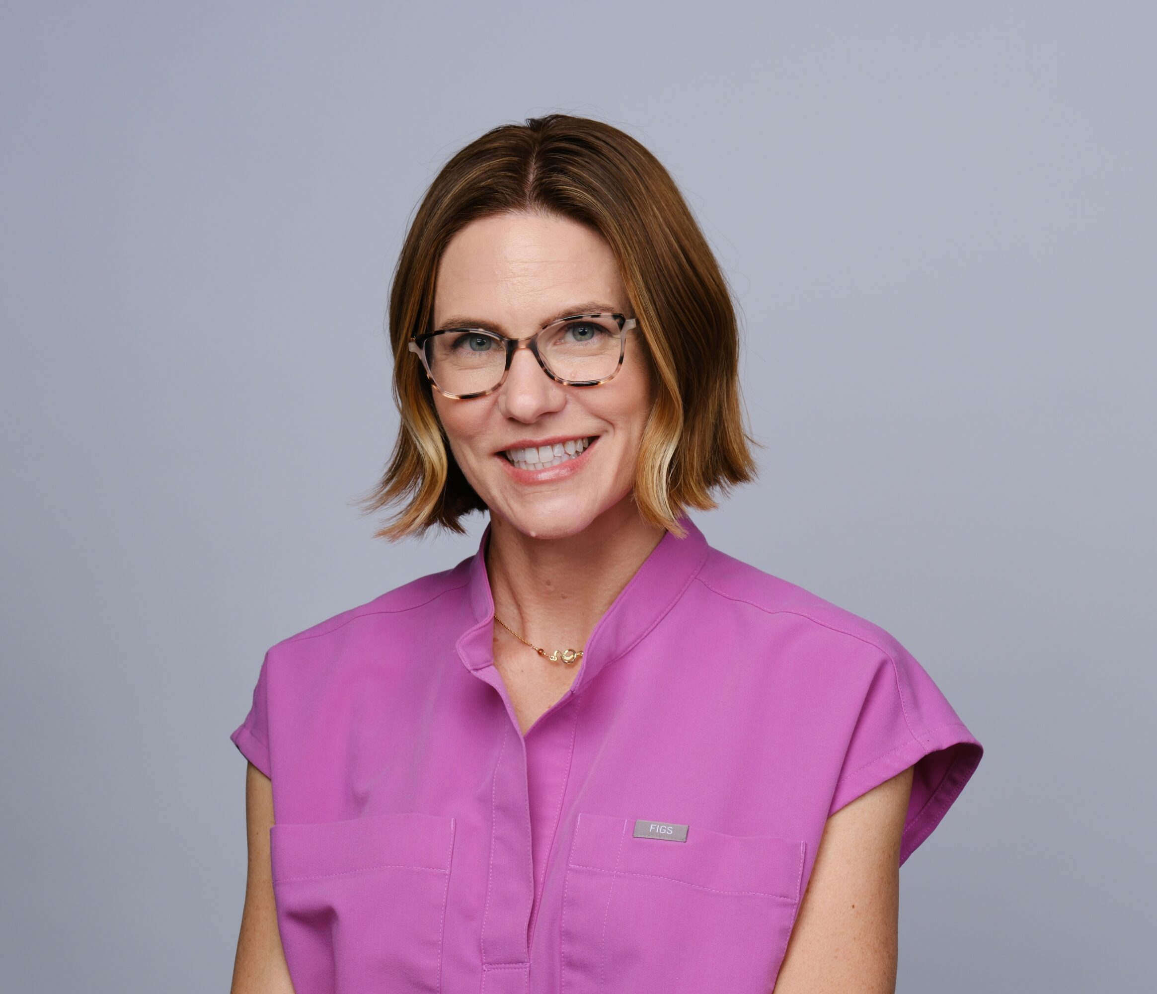 Woman with shoulder-length brown hair, glasses, smiling, wearing a pink top, against a gray background.