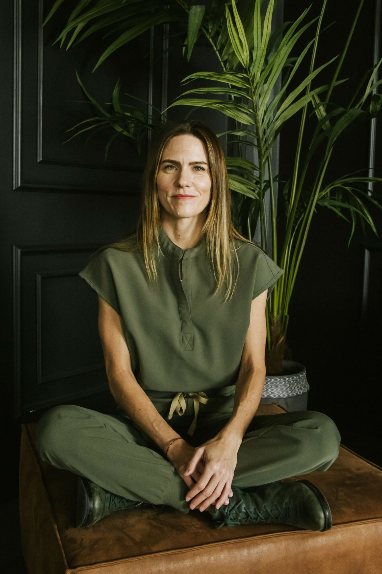 Woman sitting cross-legged on a cushion in front of a large plant and dark wall, smiling softly.