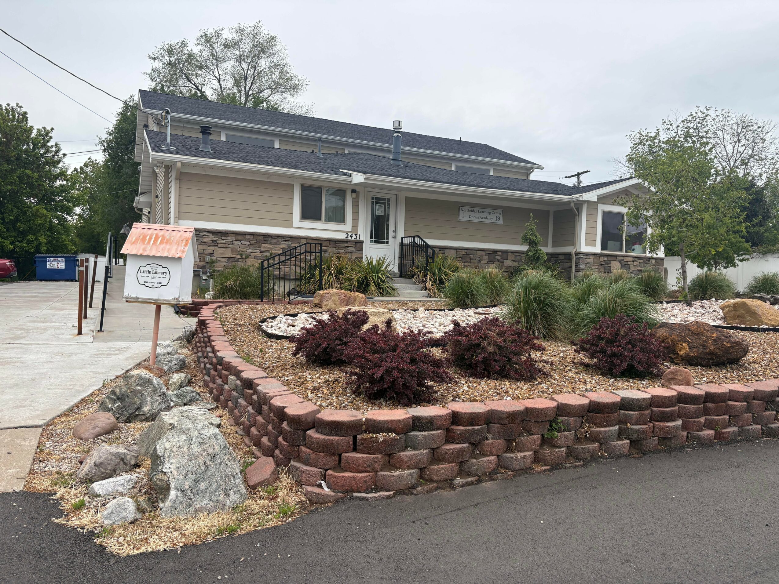 Single-story house with front porch, landscaped yard with bushes, brick border, and rocks, overcast sky.