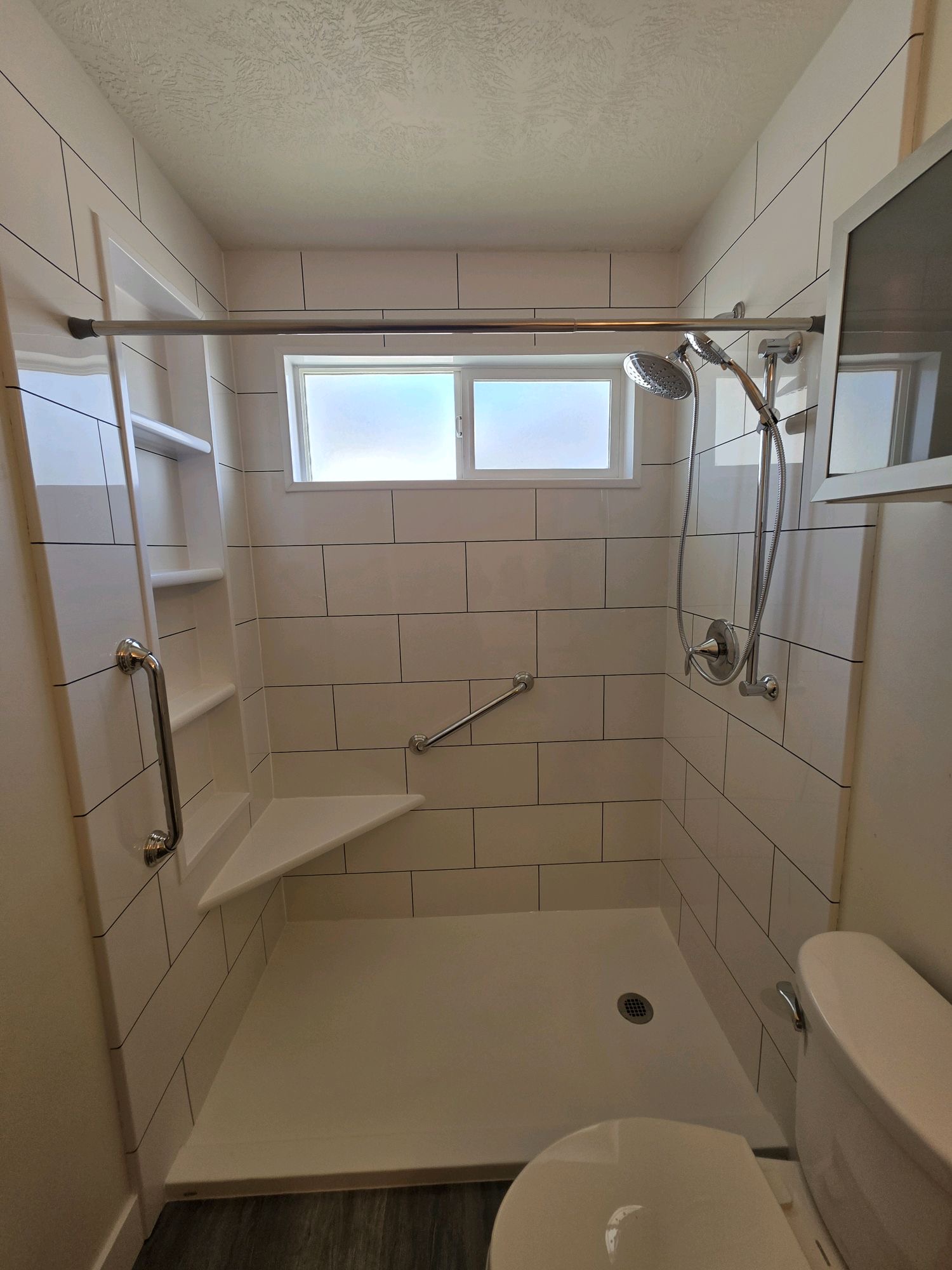Bathroom shower area with window, built-in shelves, showerhead, and grab bar, with toilet visible in foreground.