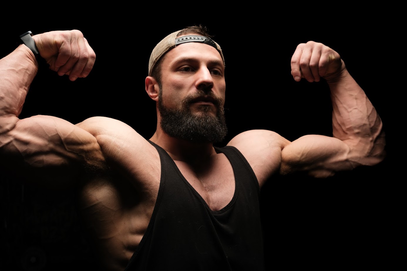 Man with a beard and cap flexing muscles, black background, wearing a sleeveless shirt.