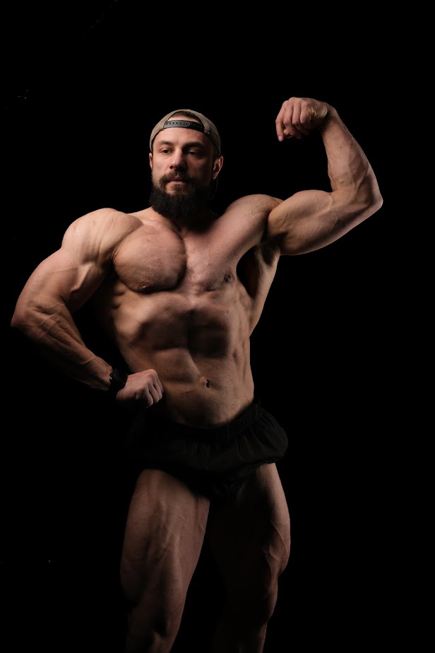 Muscular man with beard flexing arm, wearing a cap and shorts, against black background.