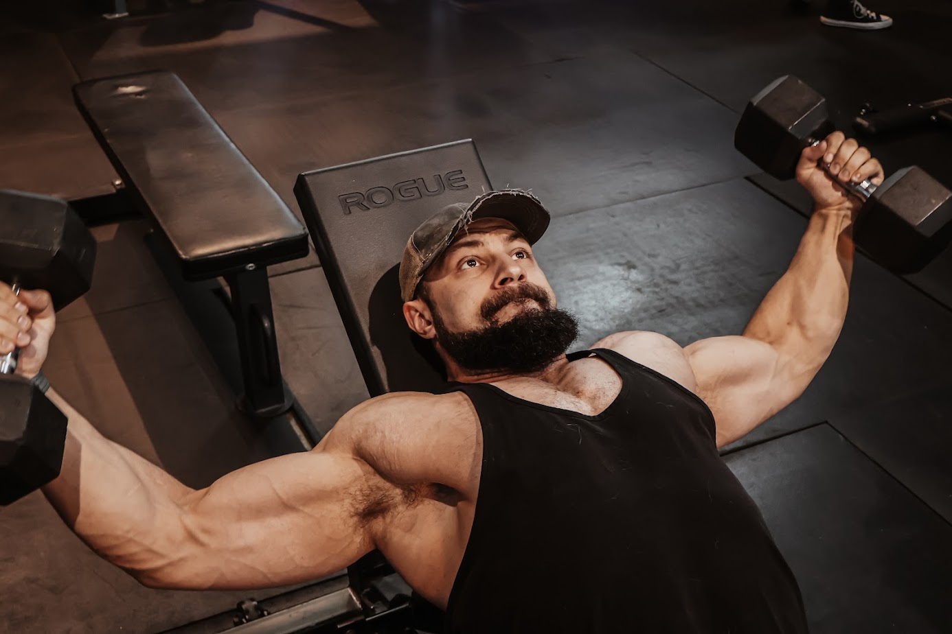 Man with a beard and cap lying on a bench, lifting dumbbells in a gym, muscles flexed, focused expression.