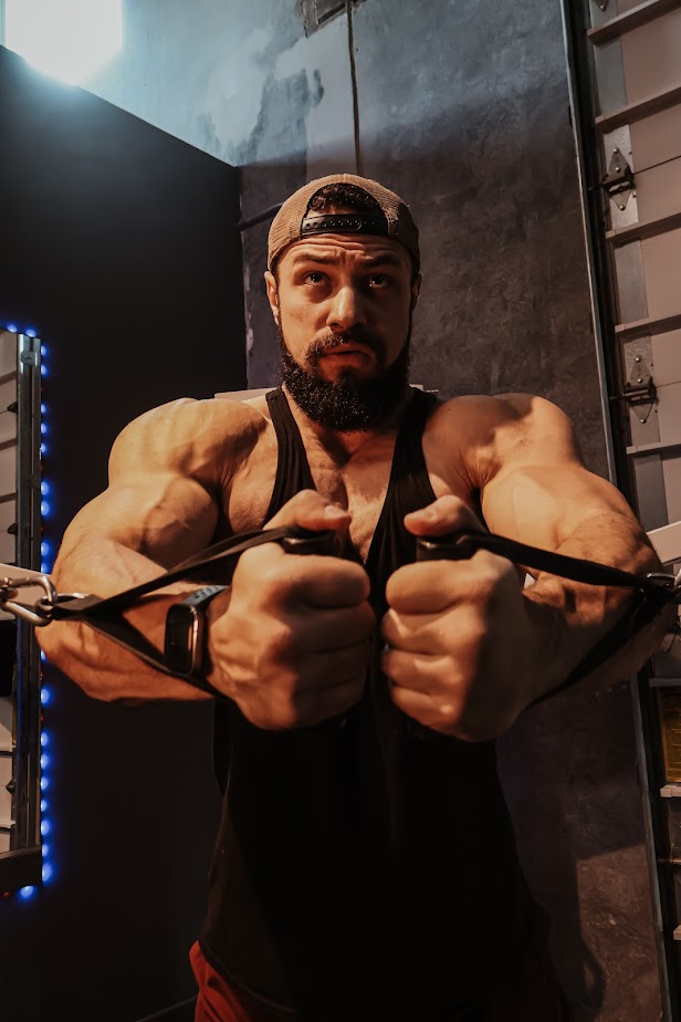 Man with a beard and cap performs a resistance exercise in a gym, showing muscular arms and focused expression.