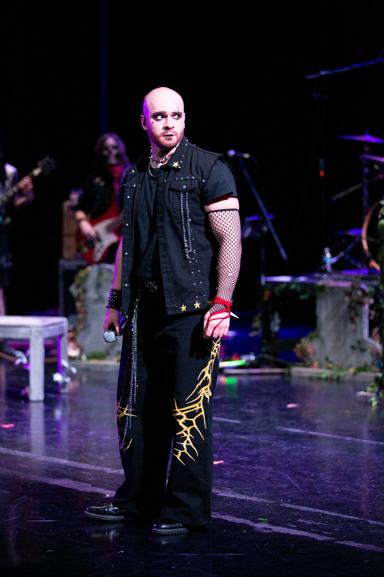 Person with a shaved head wearing a black vest with mesh sleeves, standing on stage with musical instruments in background.