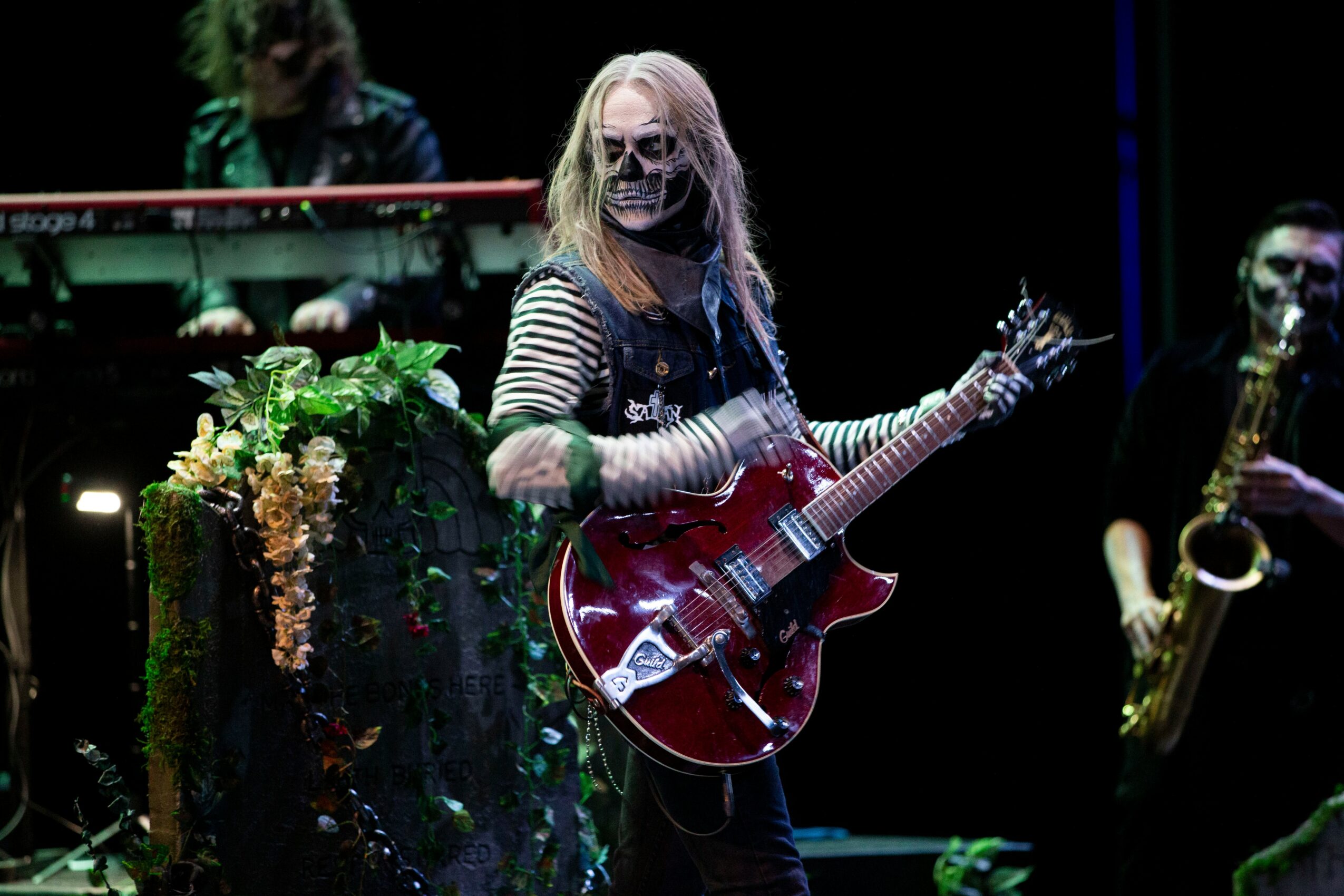 Musician with long hair, striped shirt, and face mask playing a red electric guitar on stage with a saxophonist nearby.
