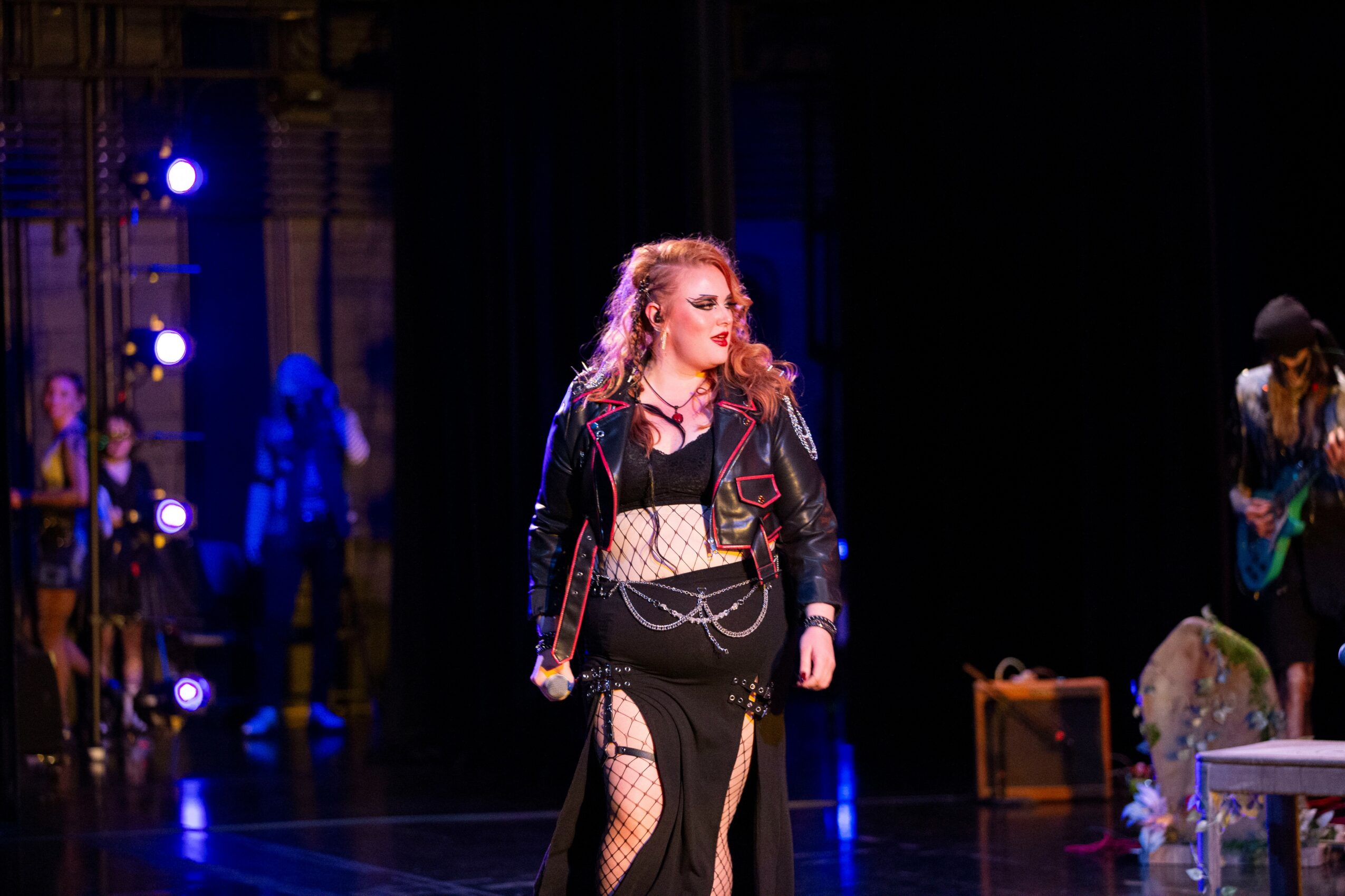 A woman with curly hair in a black outfit and leather jacket stands on stage with a dark background.