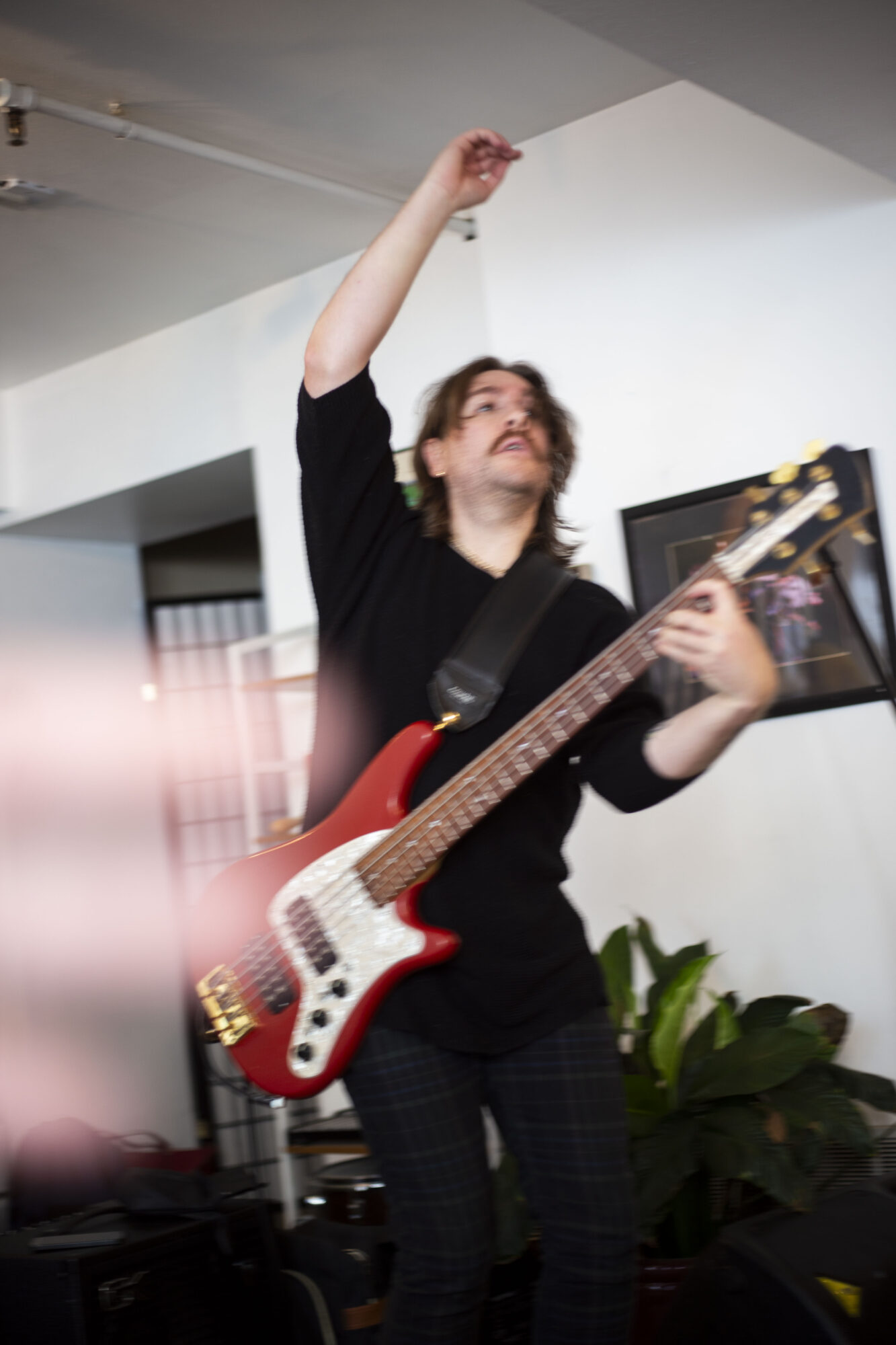 Person playing red electric guitar with one arm raised, indoors with white walls and a plant nearby.