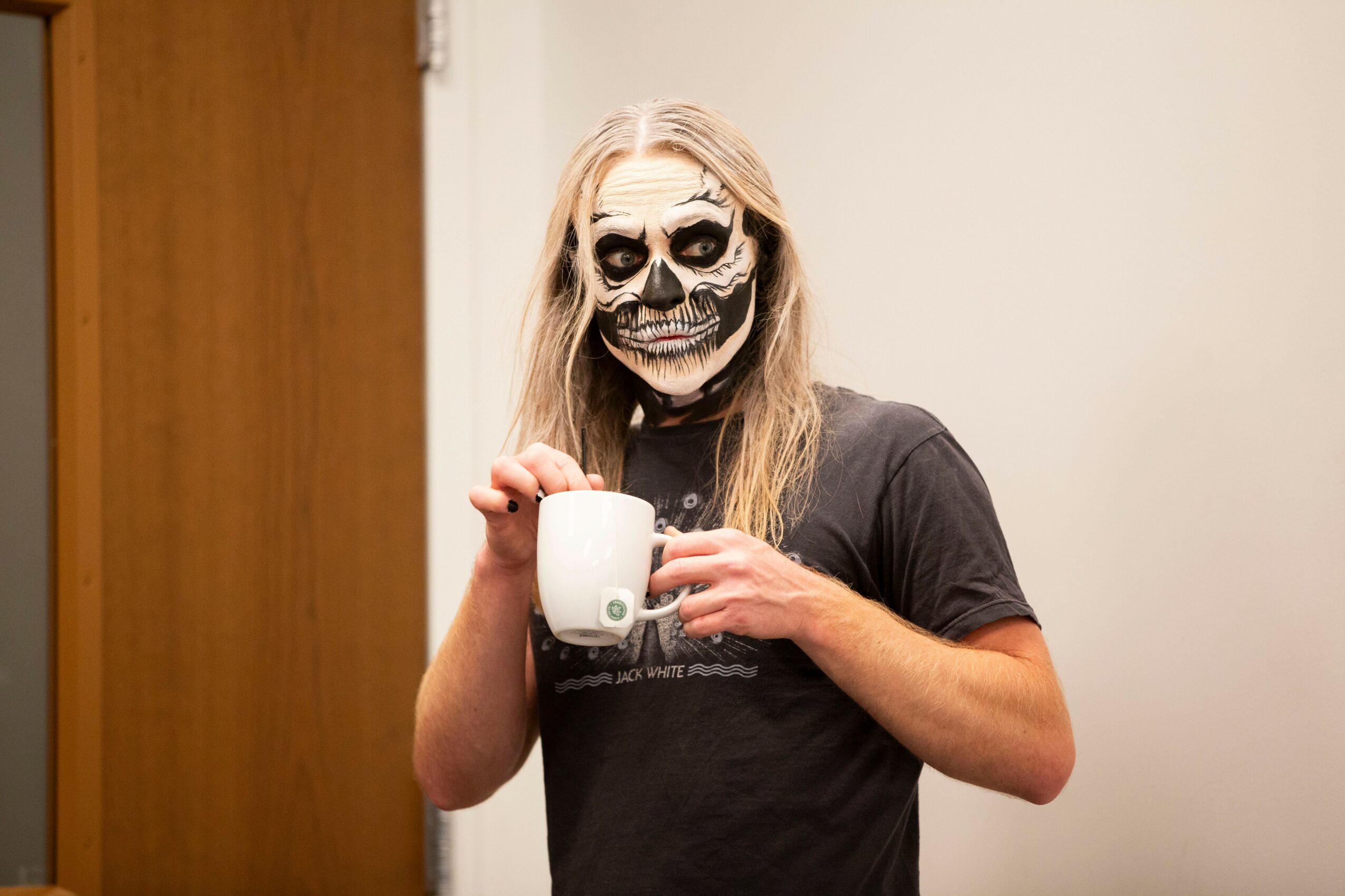 Person with skull face paint holding a white mug, standing indoors against a plain wall.