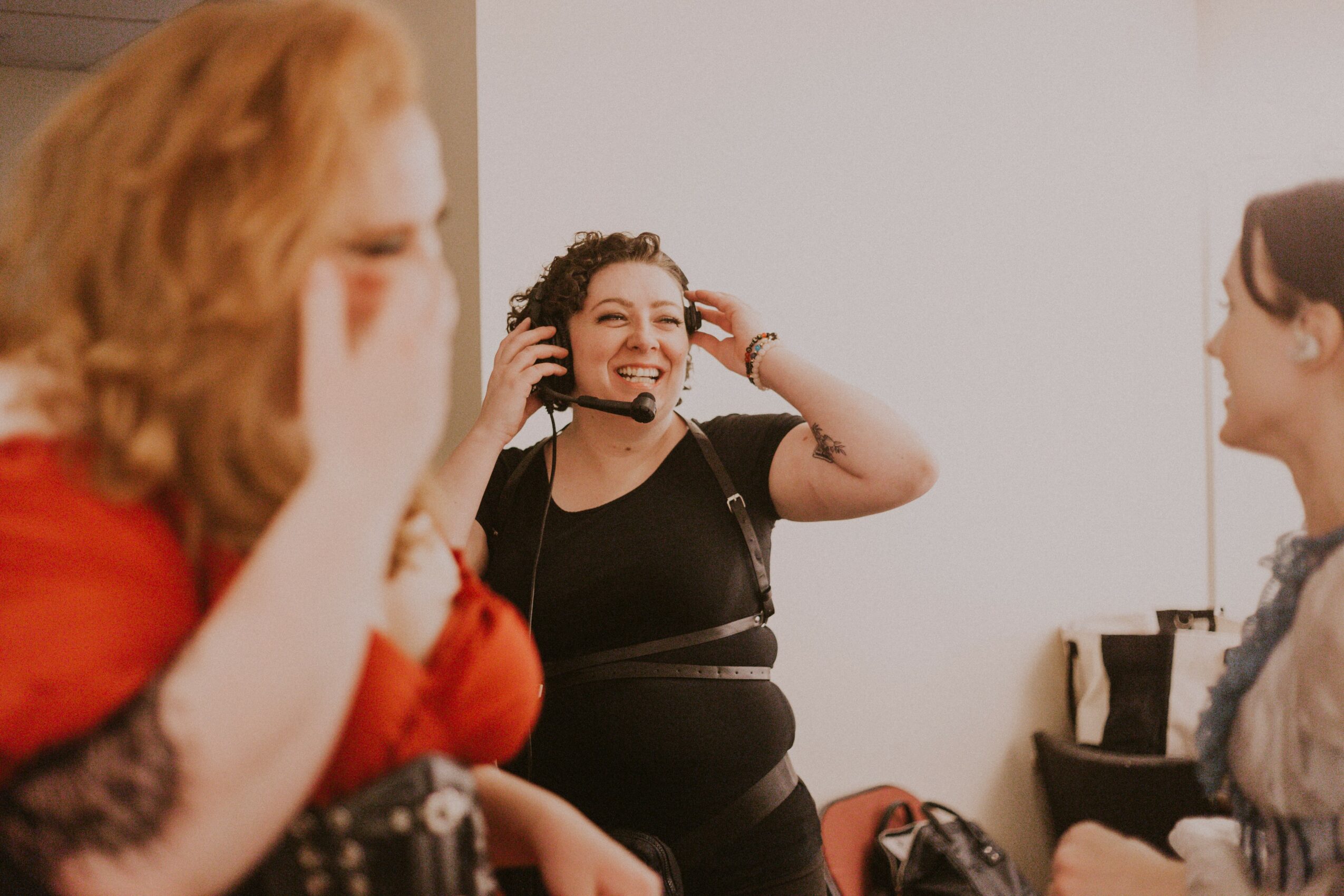 Three women in a room, one wearing a headset, smiling and laughing, with a white wall background.
