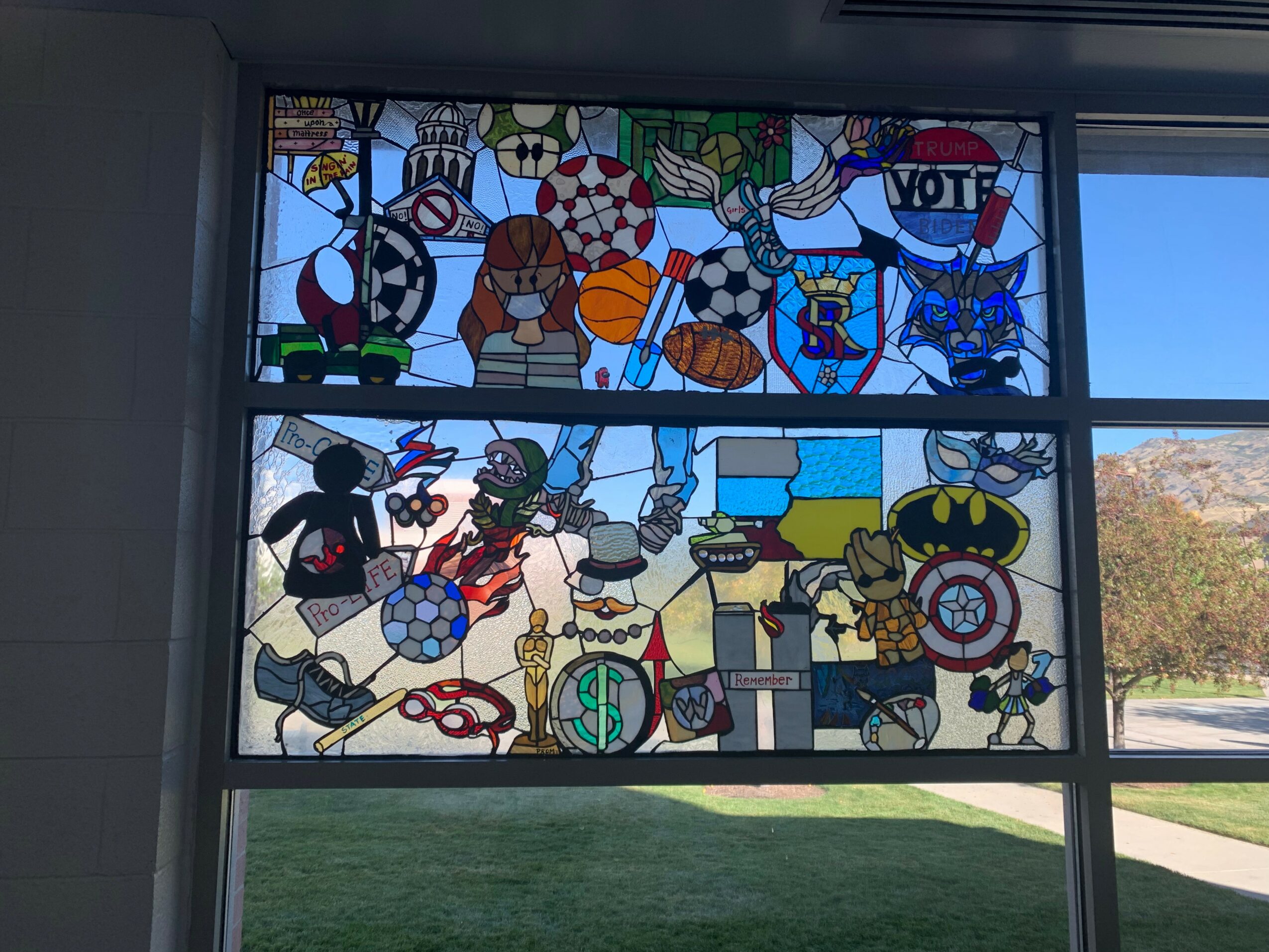 Colorful stained glass window with various sports equipment and a 'VOTE' sign, outdoors scene visible through the window.